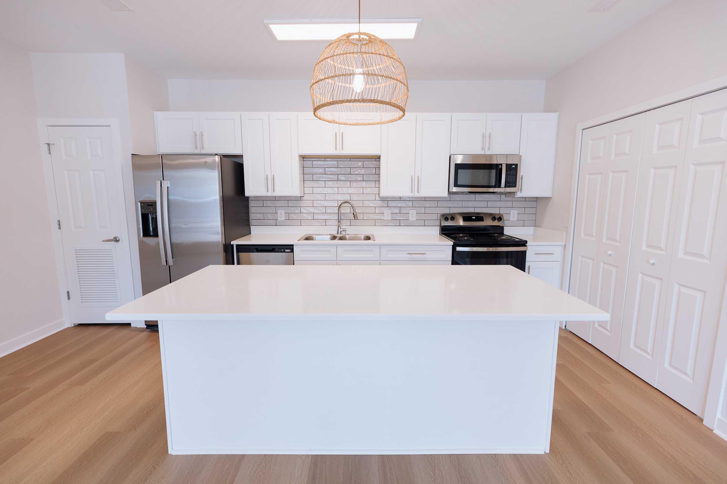 Modern kitchen featuring a large white island with a pendant light overhead. It has white cabinetry, stainless steel appliances including a refrigerator and an oven, a gray tiled backsplash, and light wood flooring, creating a bright and airy atmosphere.