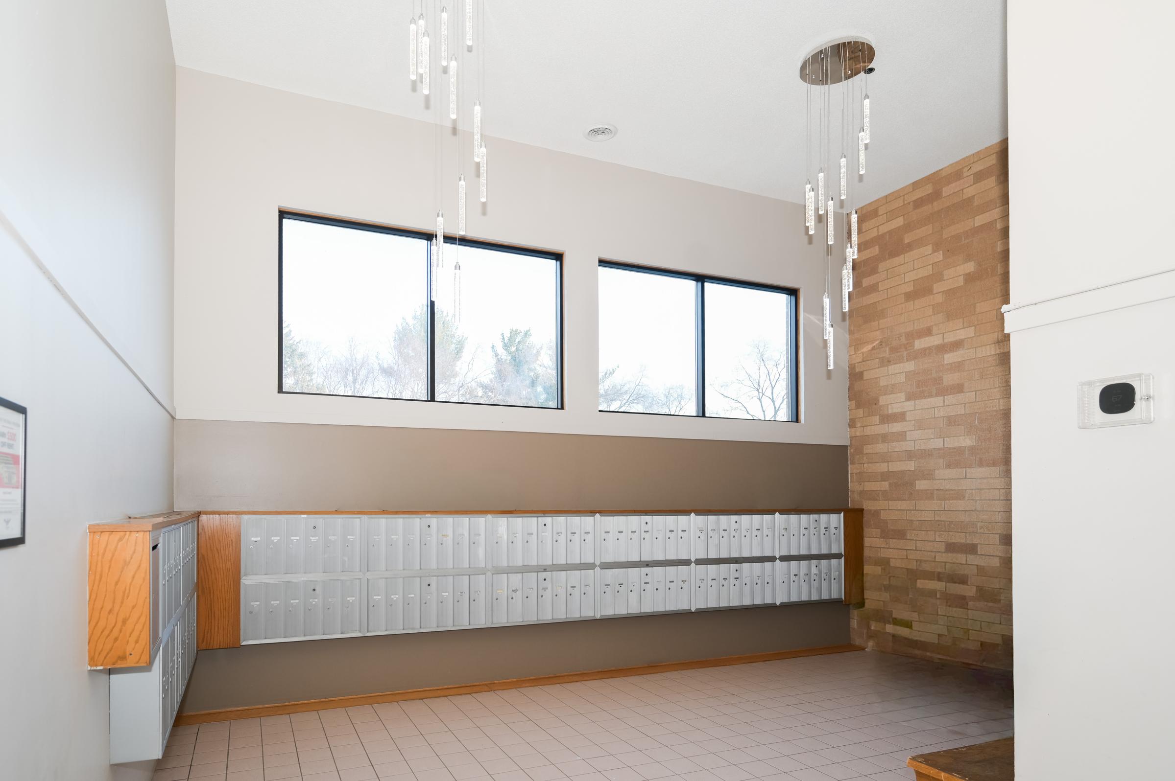 A modern lobby with wall-mounted mailboxes on the left, large windows letting in natural light, and stylish hanging light fixtures from the ceiling. The walls feature a combination of light colors and brick, creating a clean and inviting atmosphere.