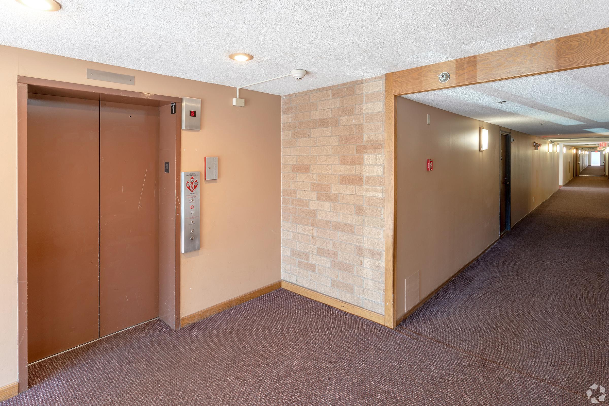 A hallway featuring an elevator. The walls are painted a light color, and there's a textured section made of brick. The carpet is dark, and doors line the corridor. The elevator is situated on the left side, marked with buttons and a floor indicator.