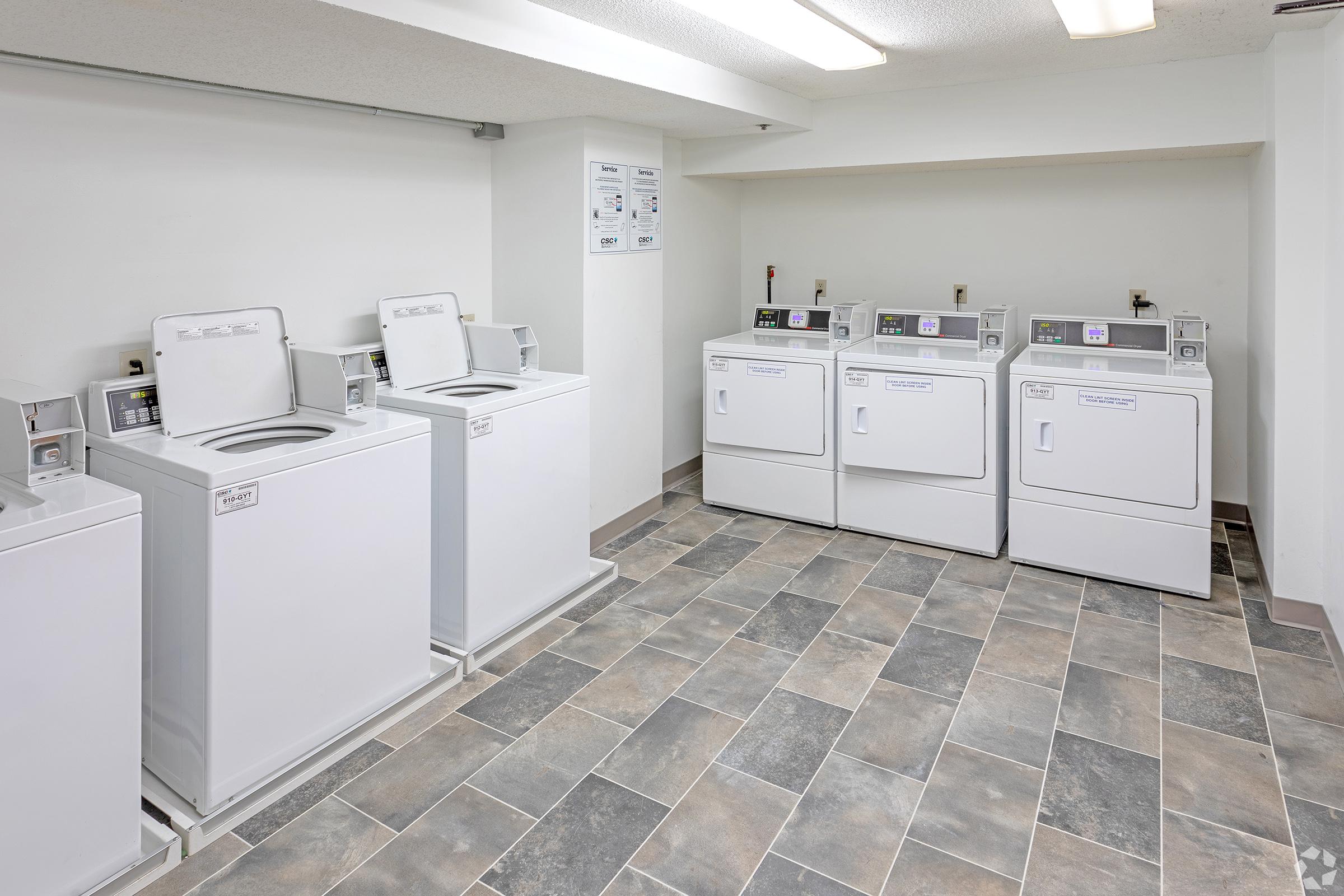 A clean and modern laundry room featuring several white top-loading washing machines and front-loading dryers arranged in a spacious, well-lit area. The room has grey and beige tiled flooring and white walls, creating a bright and inviting atmosphere.