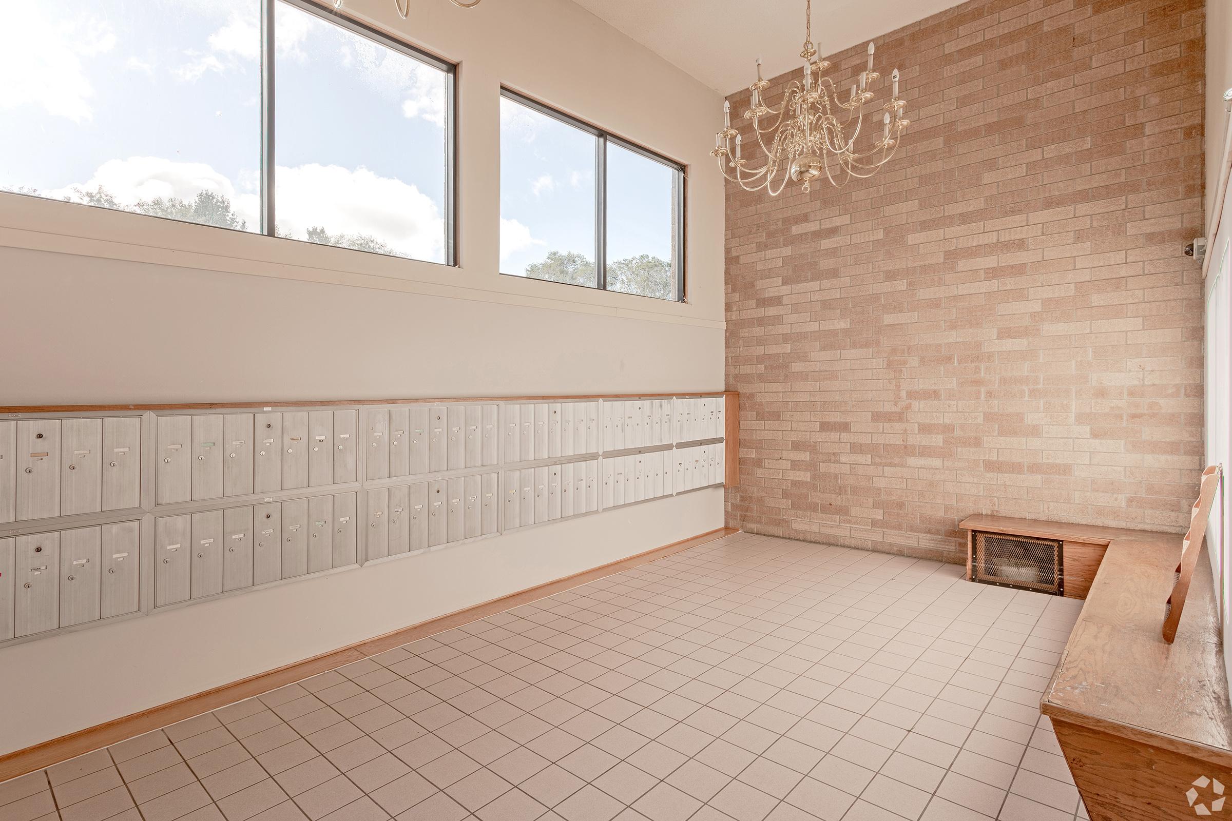 A well-lit mailroom featuring a row of silver mailboxes on the left, a wooden bench along the wall, and large windows allowing natural light. The right wall has a textured brick design and a decorative chandelier hanging from the ceiling.