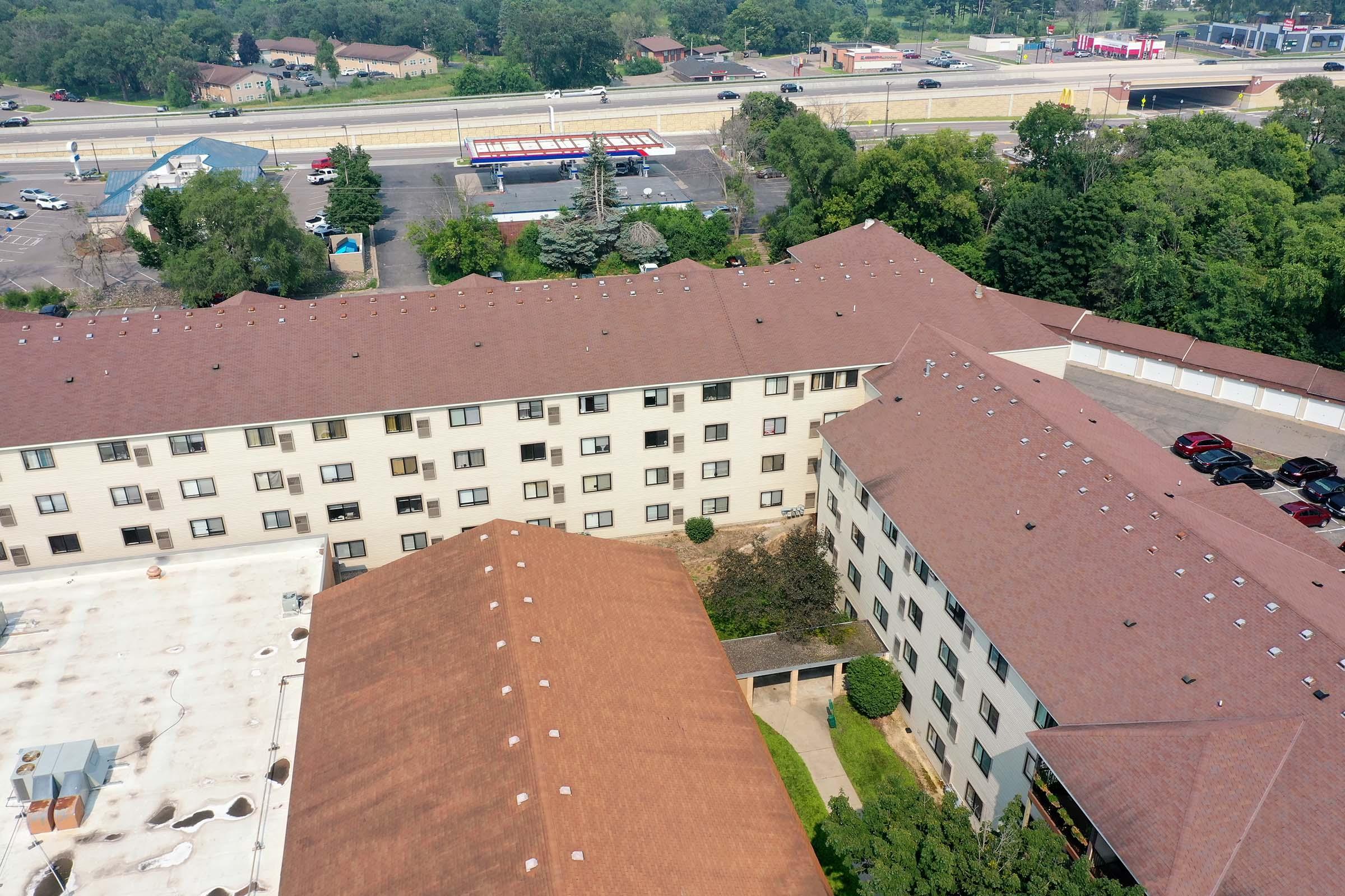 Aerial view of two adjacent multi-story buildings with flat roofs and multiple windows. One building is topped with a red roof, featuring parking areas nearby. The background shows a highway with vehicles and some greenery.