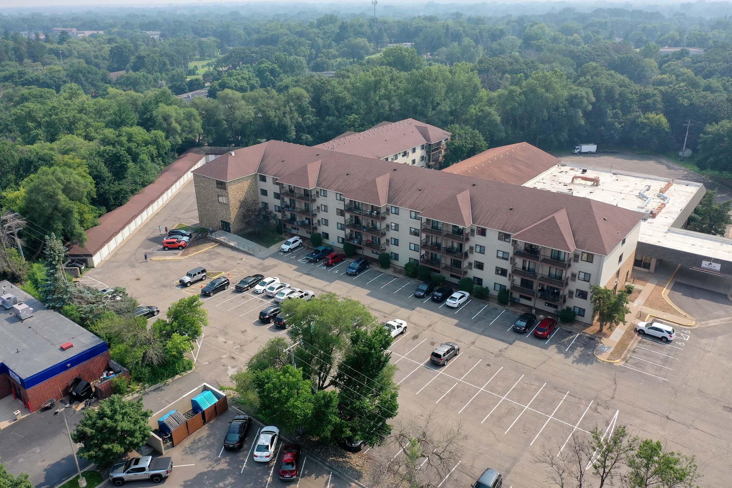 Aerial view of a multi-story residential building surrounded by trees, with a parking lot in front. Several cars are parked in designated spaces, with some empty spots visible. The area is green, indicating a suburban setting with lush vegetation.