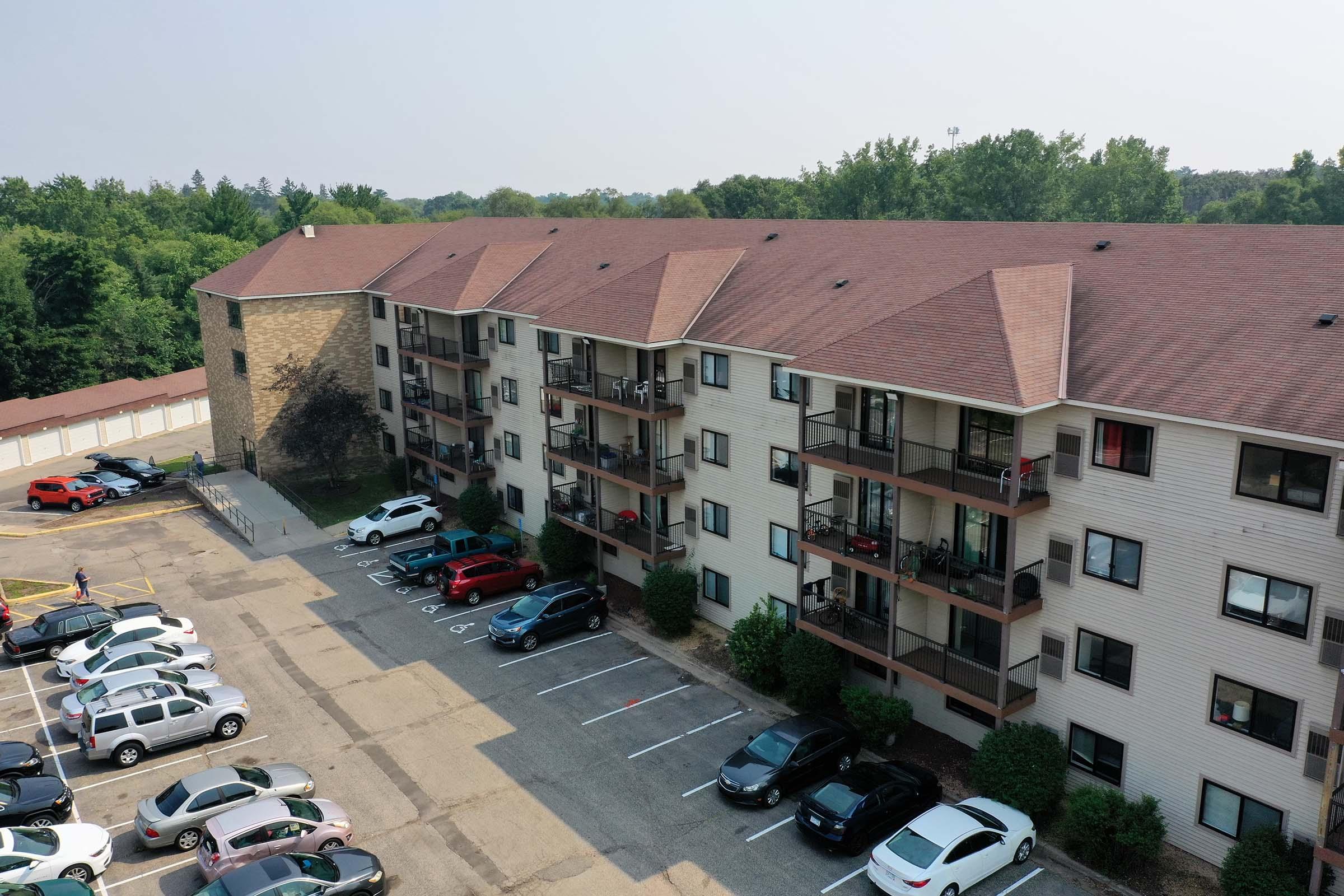 Aerial view of a multi-story apartment building with balconies, surrounded by green trees. Several parked cars are visible in the foreground, with a small section of pavement. The building features a light-colored exterior and red roof, with a few residents visible on the balconies.