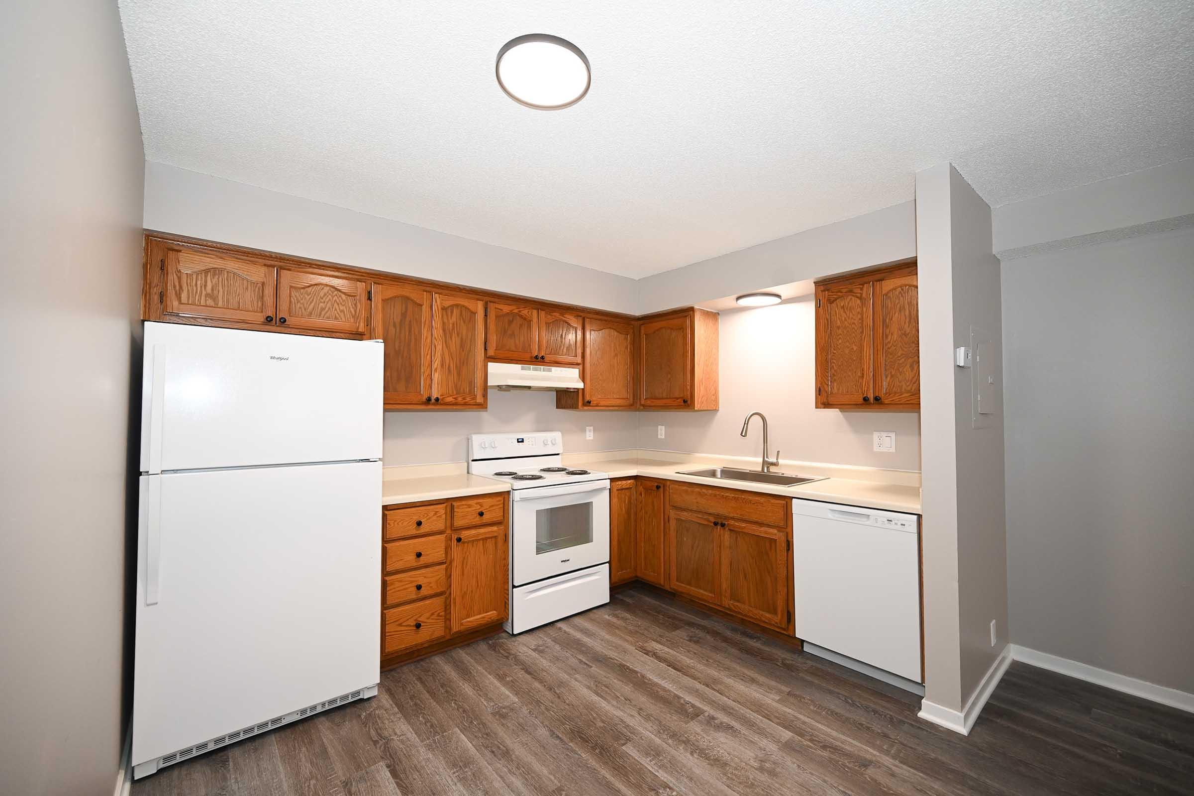 Modern kitchen with wooden cabinets, white appliances including a refrigerator, stove, and dishwasher. The countertop is light-colored with a sink underneath a circular overhead light. The floor features dark wood-like vinyl, and the walls are painted in a neutral tone.