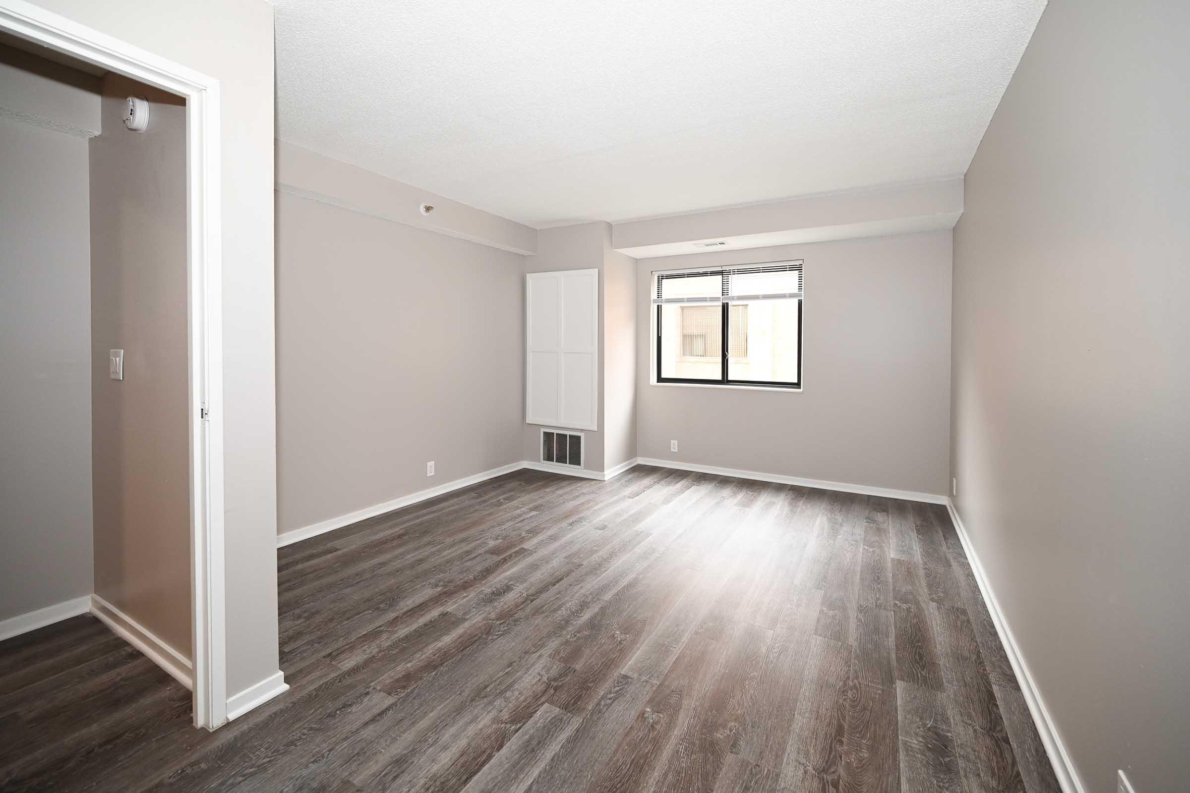 A spacious, empty room with light gray walls and new flooring. The floor is made of laminate wood, and there's a large window allowing natural light. A small wall-mounted shelf is visible, and one corner shows a door leading to another area. This space is ready for personalization and decoration.