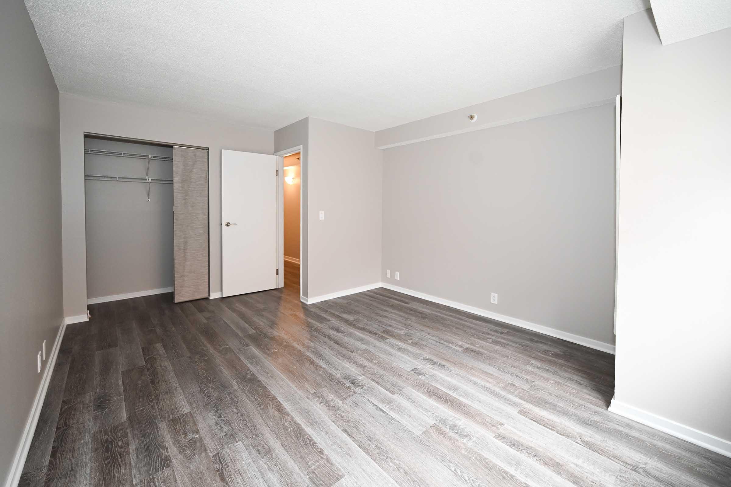 Empty room with light gray walls and wood-like flooring. An open closet is visible on the left, and a doorway leads to another room on the right. Natural light is coming from the ceiling light fixture. The space appears modern and freshly painted, suitable for various uses.