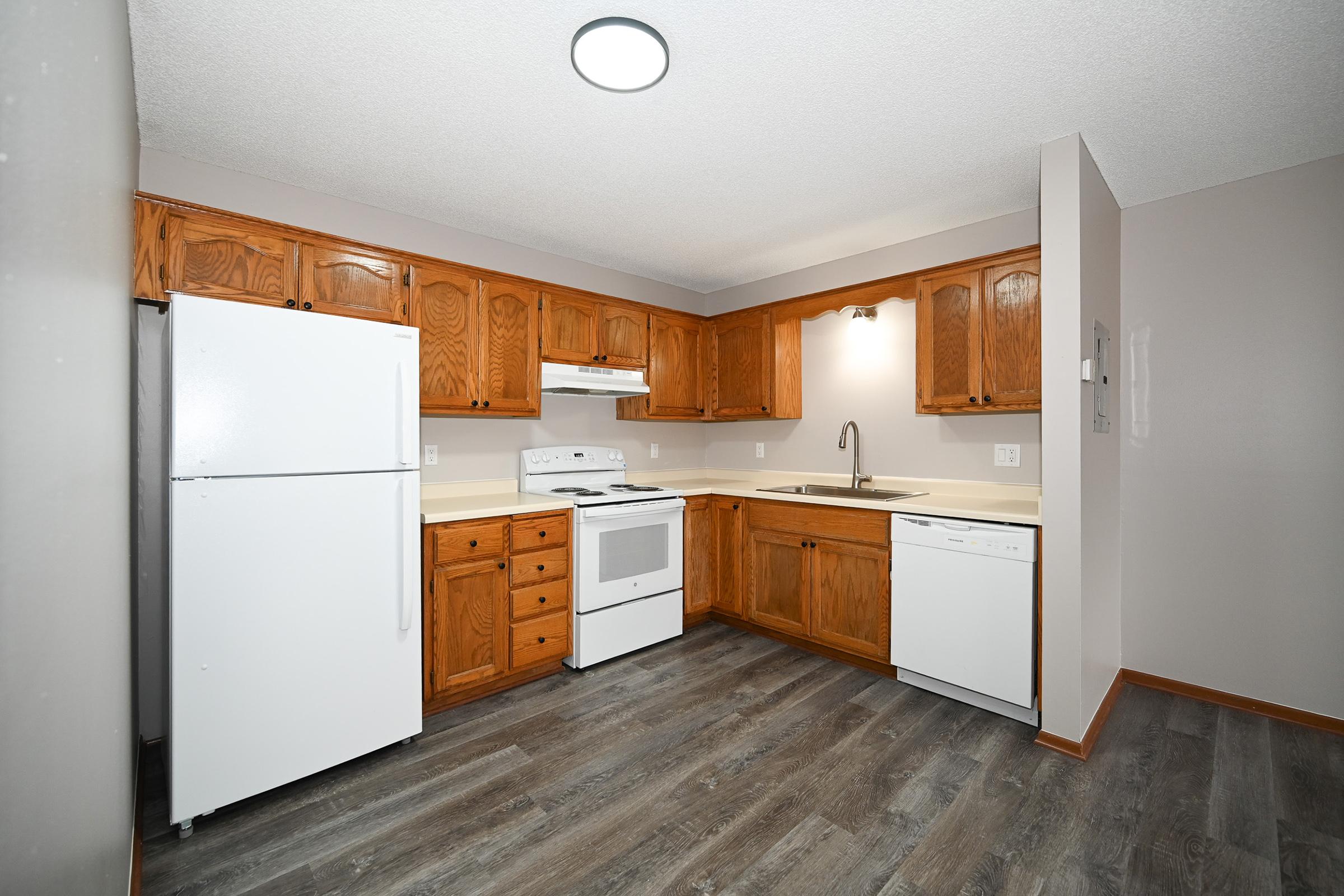 A modern kitchen featuring wooden cabinets, a white refrigerator, a stove, and a dishwasher. The countertop is light-colored, and there's a sink under a window. The flooring is a dark laminate, and the overall layout is open and spacious with ceiling lights overhead.