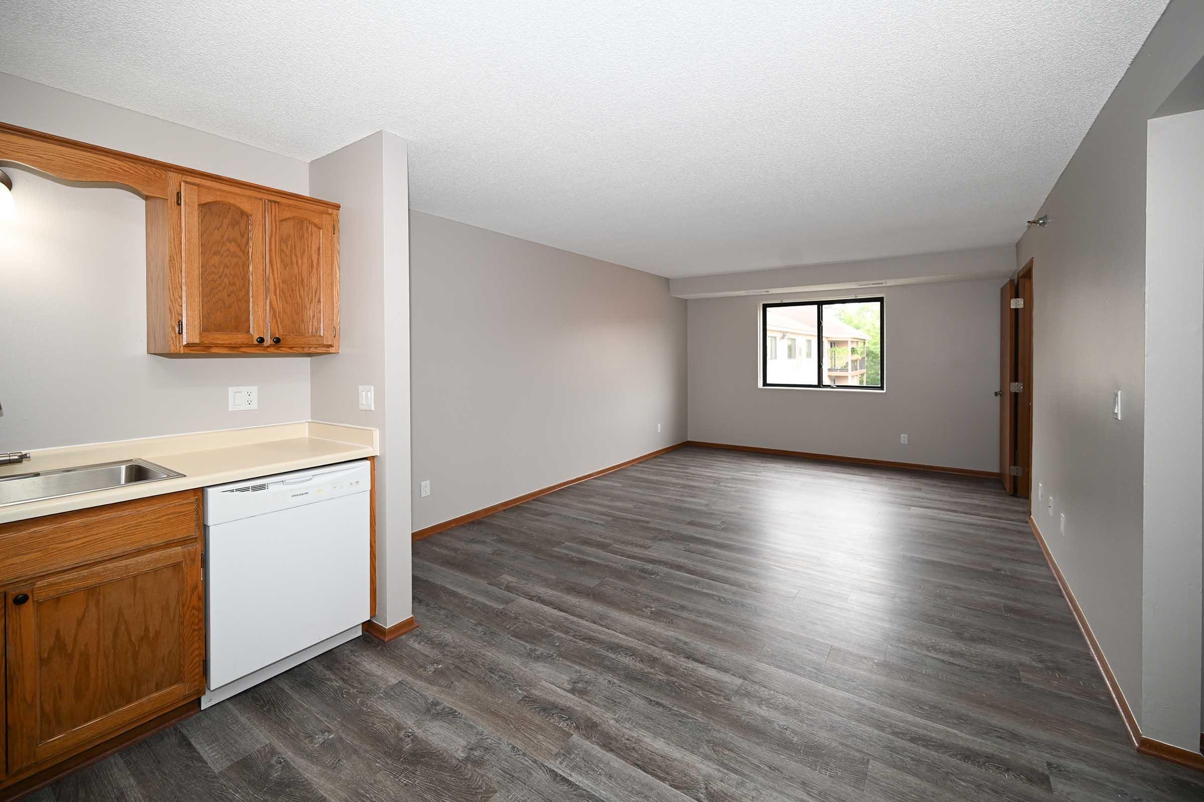 Interior view of a spacious, empty apartment featuring light wood cabinetry in the kitchen area, a white dishwasher, and a large window providing natural light. The flooring is dark wood laminate, and walls are painted a neutral gray, creating a bright, inviting atmosphere.