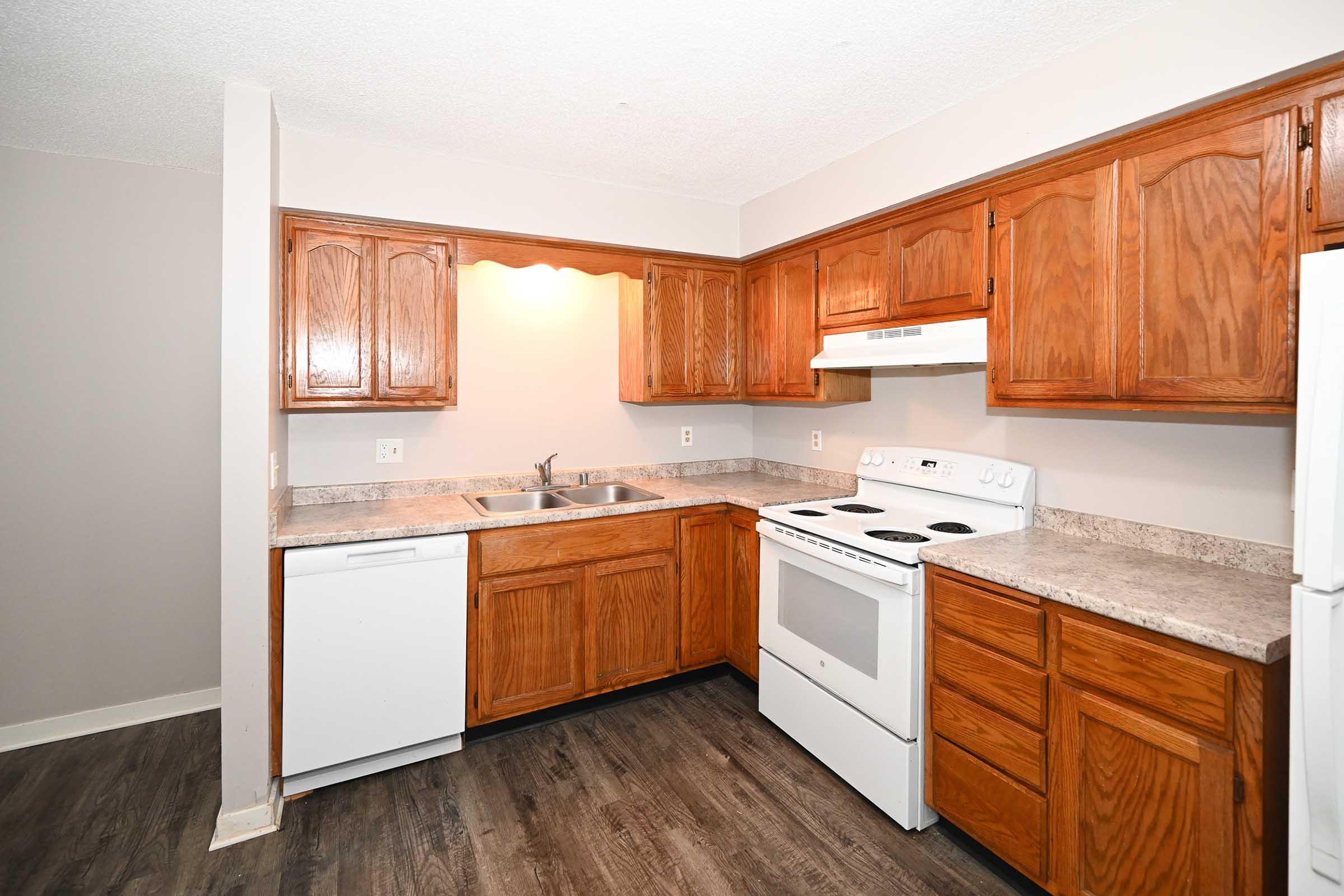 A modern kitchen featuring wooden cabinets, a double sink, a white stove, and a dishwasher. The countertops are light-colored, and the floor has a dark wood-like finish. The walls are painted in a neutral tone, creating a warm and inviting atmosphere.