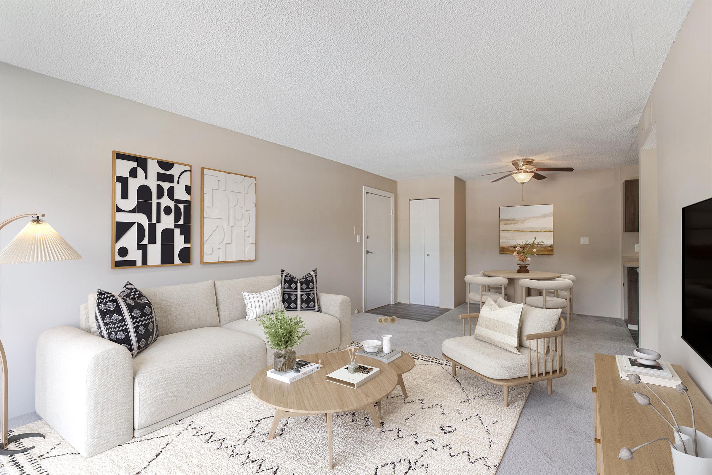 A stylish living room featuring a light-colored sofa with black and white accent pillows, a round wooden coffee table, and a light rug. There's a dining area with a circular table and chairs, and framed artwork on the wall. A ceiling fan and television are visible, creating a cozy, modern atmosphere.