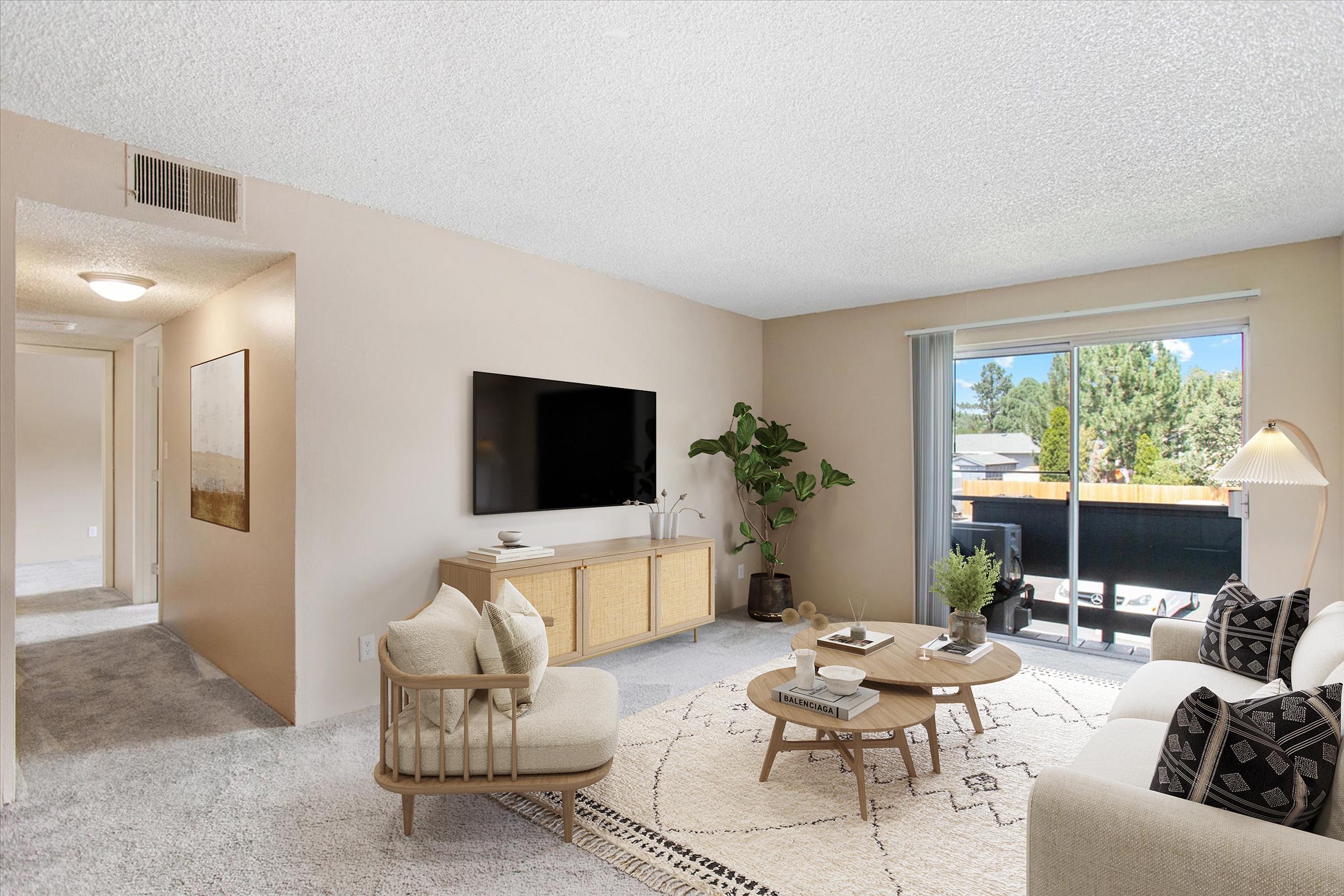 A cozy living room featuring a light-colored sofa, a circular coffee table, and a large TV mounted on a wooden console. There is a potted plant in the corner and decorative items on the coffee table. Natural light floods in from a nearby window, enhancing the warm and inviting atmosphere.