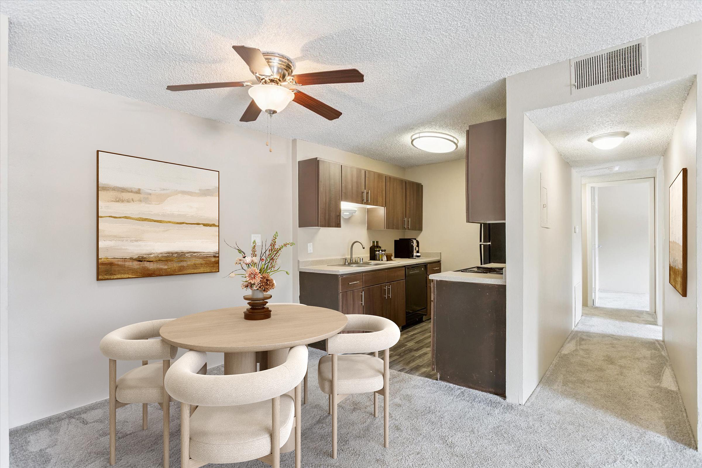 A modern kitchen and dining area featuring a round wooden table with four beige chairs, a ceiling fan, and a dark cabinetry kitchen. The walls are light-colored, and a hallway leading to other rooms is visible in the background. A framed painting is displayed on the wall.