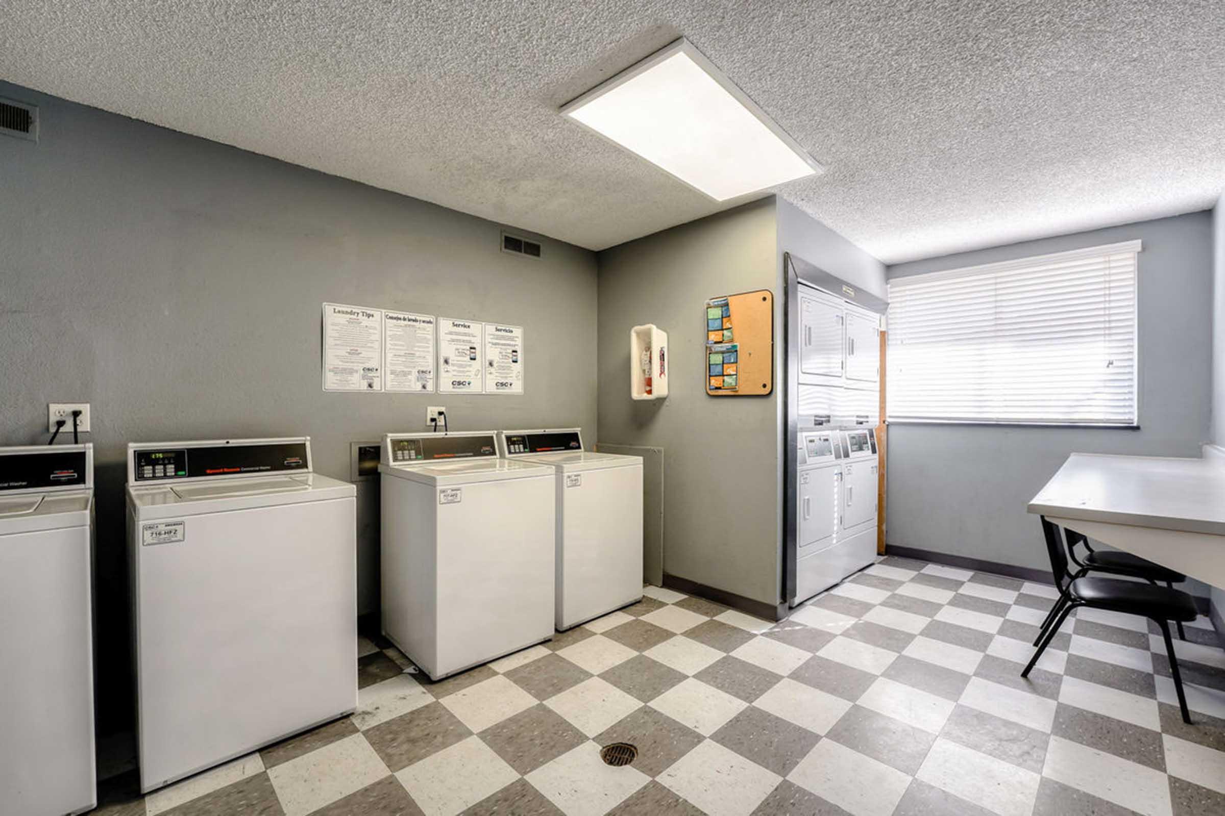 A clean laundry room featuring several white washing machines and dryers lined up against a gray wall. The floor has a checkerboard pattern in gray and white tiles. A large window provides natural light, and a table with a chair is positioned nearby. Noticeable instruction signs are posted on the wall.