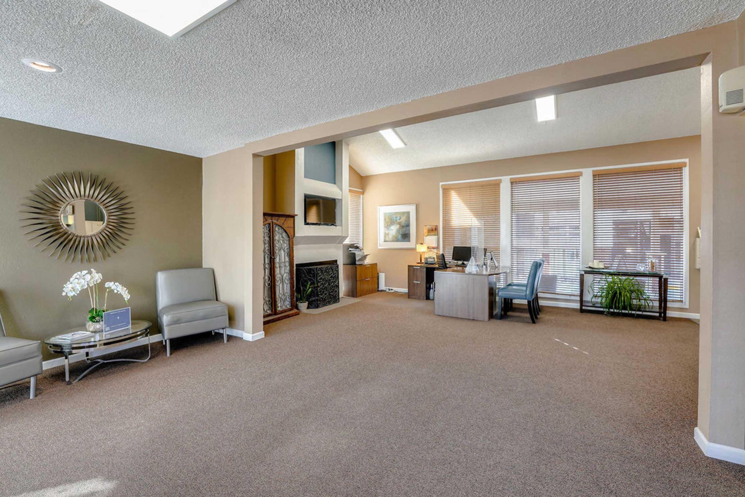 Interior view of a modern office space featuring a cozy seating area with two gray chairs, a small side table, and decorative elements like an ornamental mirror. A desk is positioned nearby, along with large windows allowing natural light and plants for a welcoming atmosphere.