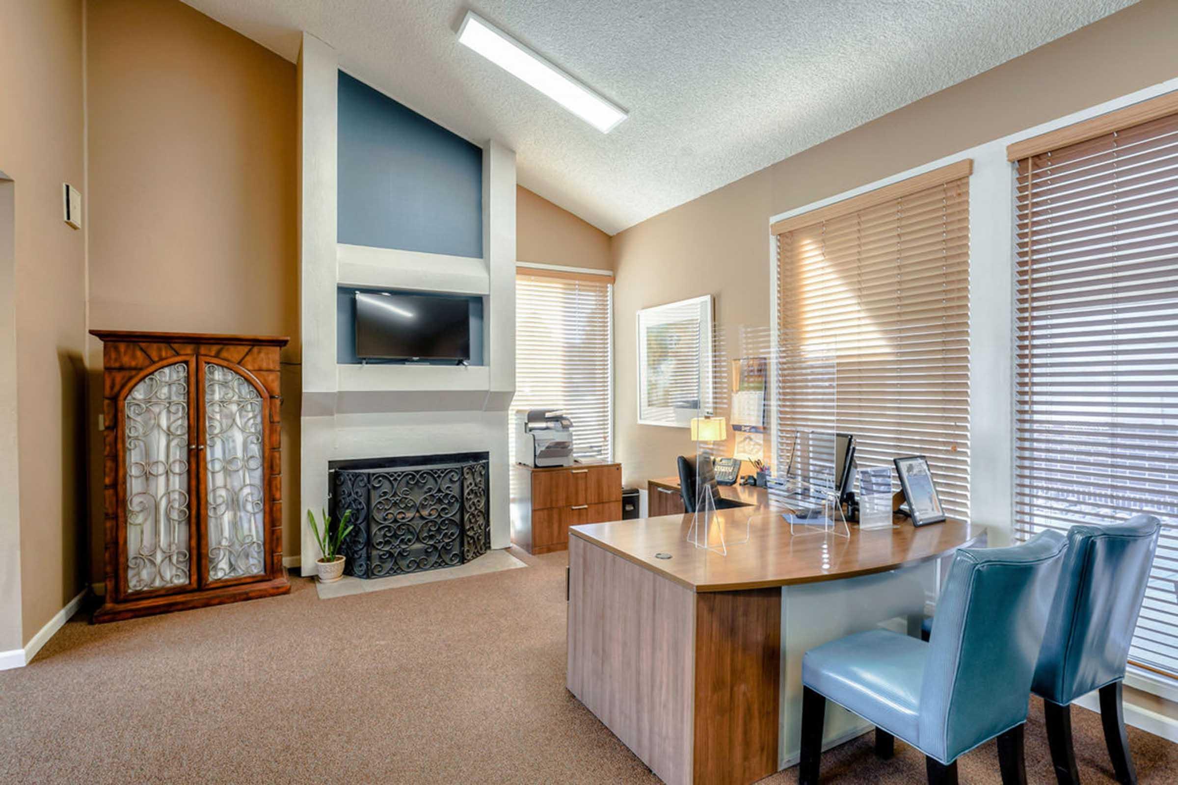 A cozy home office featuring a wooden desk with a blue chair, a modern wall-mounted TV, and a decorative fireplace. Natural light floods the room through wooden blinds, illuminating a printer and framed pictures on the desk. A stylish cabinet and a potted plant add to the inviting atmosphere.