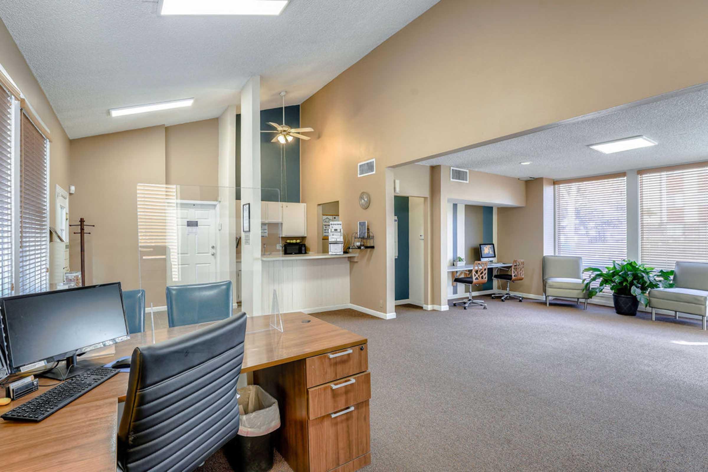 A modern office space featuring a wooden desk with a computer and a chair, set in a room with beige walls and carpet. A small seating area with green chairs and plants is visible. A kitchenette area is in the background, along with a ceiling fan and natural light from windows.