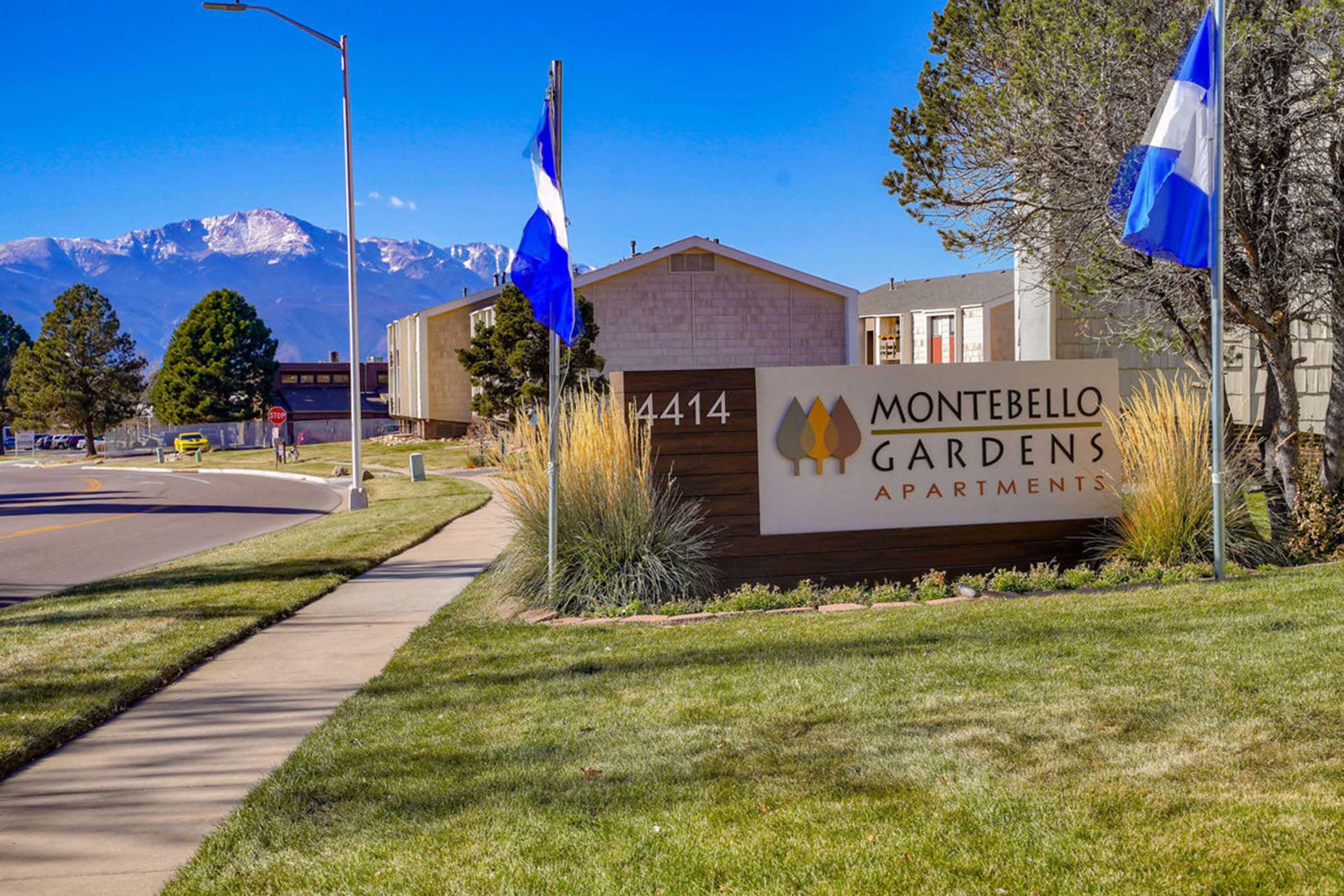 Sign for Montebello Gardens Apartments with blue flags and a landscaped area. In the background, mountains are visible, contributing to a scenic view. The setting is sunny with clear skies.