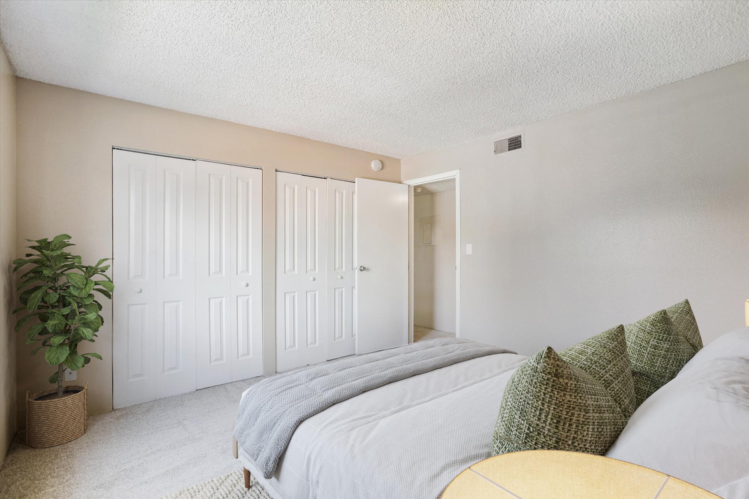 Cozy bedroom featuring a light gray bed with decorative green pillows, a small round table, and a potted plant. The room has beige walls, carpet flooring, and white double doors leading to a closet. An open door reveals a bathroom area, enhancing the spacious feel of the room.
