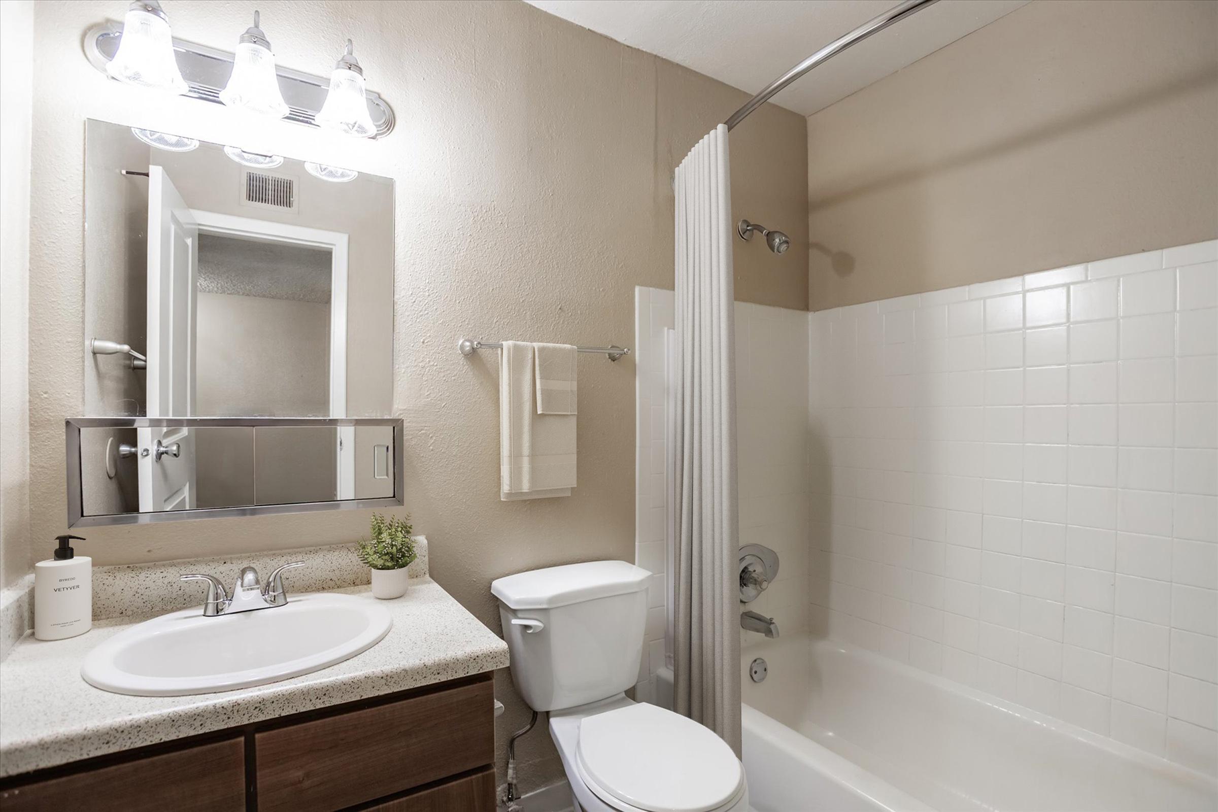 A clean, modern bathroom featuring a white bathtub with a shower curtain, a mirror above a sink with a countertop, a toilet, a towel rack, and a small potted plant. Soft lighting from a fixture above the mirror enhances the neutral color scheme of the walls.