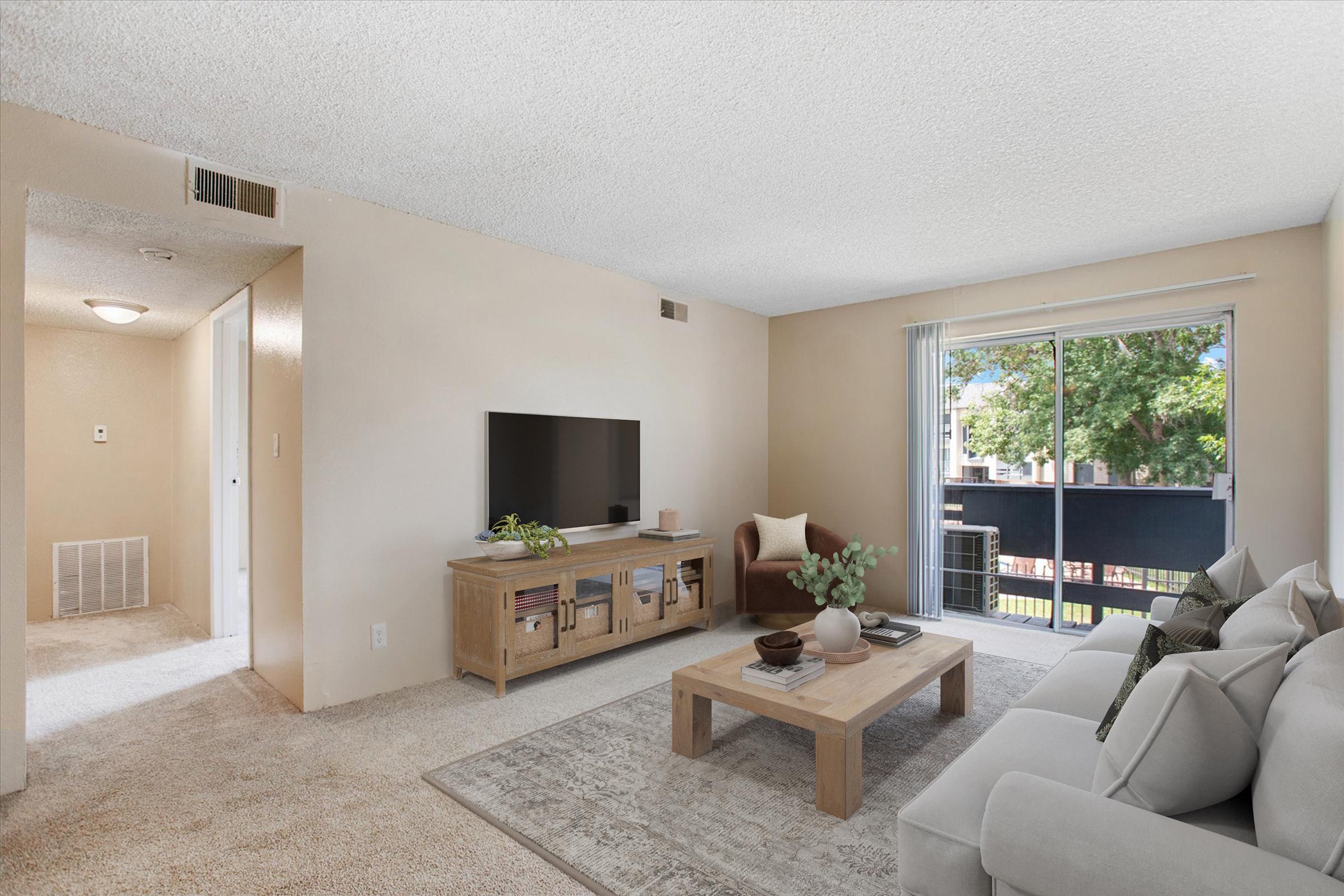 A cozy living room featuring a light-colored couch, a wooden coffee table, a television on a wood cabinet, and a potted plant. Large windows let in natural light, revealing a view of greenery outside. The walls are painted in neutral tones, and the carpet adds warmth to the space.