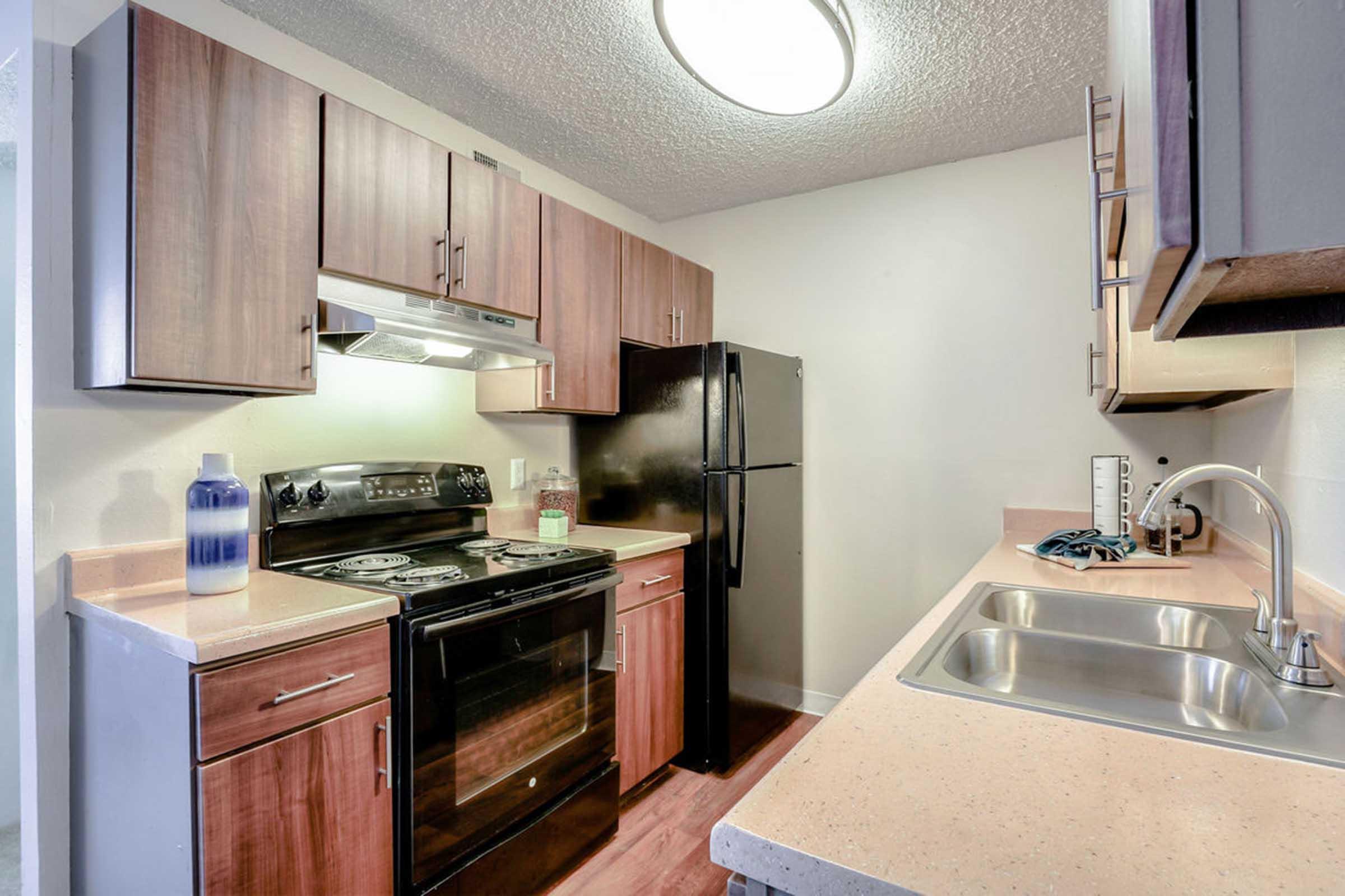 Modern kitchen featuring wooden cabinets, a black refrigerator, and a stove with an overhead microwave. The countertop is light-colored, with a double sink. A decorative blue vase is positioned next to the stove, and there are kitchen utensils on the wall. Bright ceiling light enhances the space.