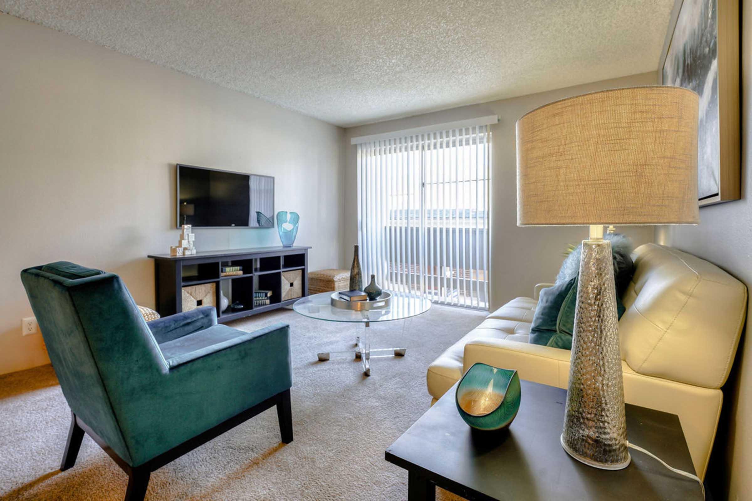 A modern living room featuring a light-colored sofa and teal accent chairs. A glass coffee table sits in the center, and there's a black entertainment unit with a TV. Natural light streams in through vertical blinds, illuminating the space. Decorative items are displayed on the coffee table and the unit.