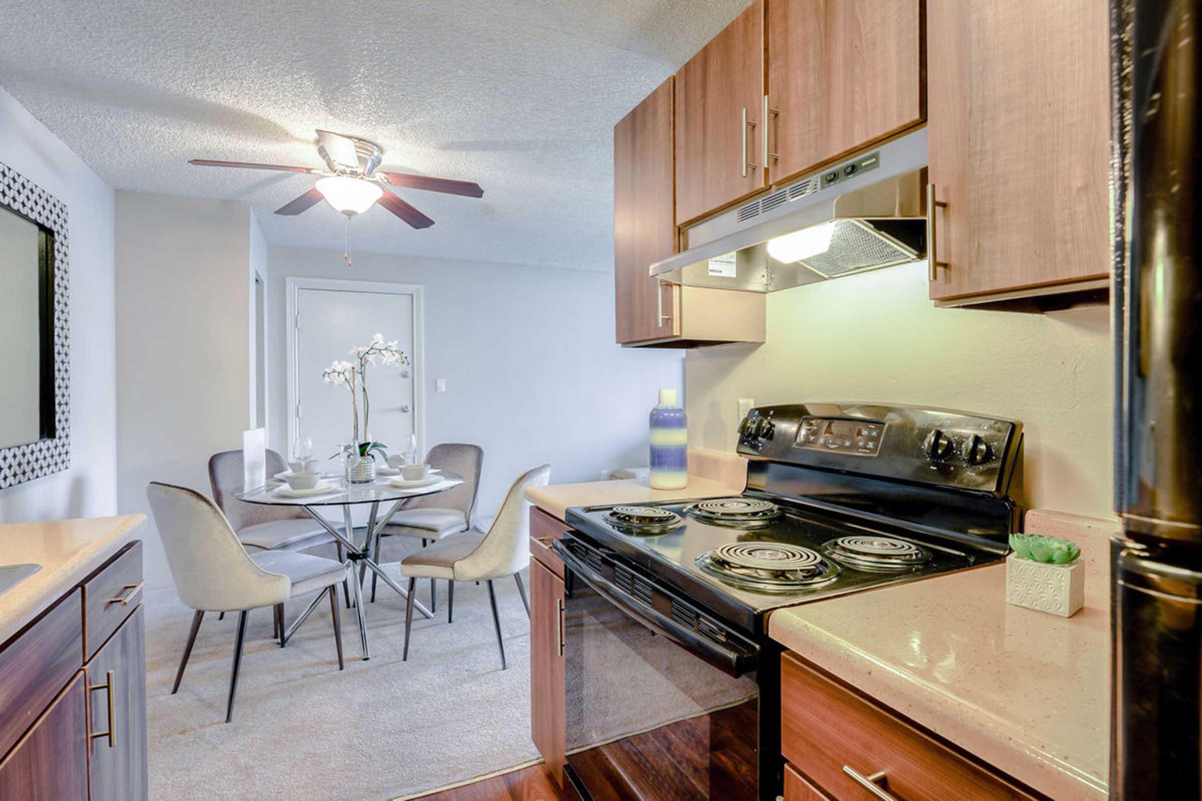 A modern kitchen featuring a stove, range hood, and cabinets in warm wood tones. Adjacent to the kitchen is a dining area with a round table and four stylish chairs. The space is illuminated by a ceiling fan, and there are decorative elements, including a small plant and a vase, enhancing the ambiance.