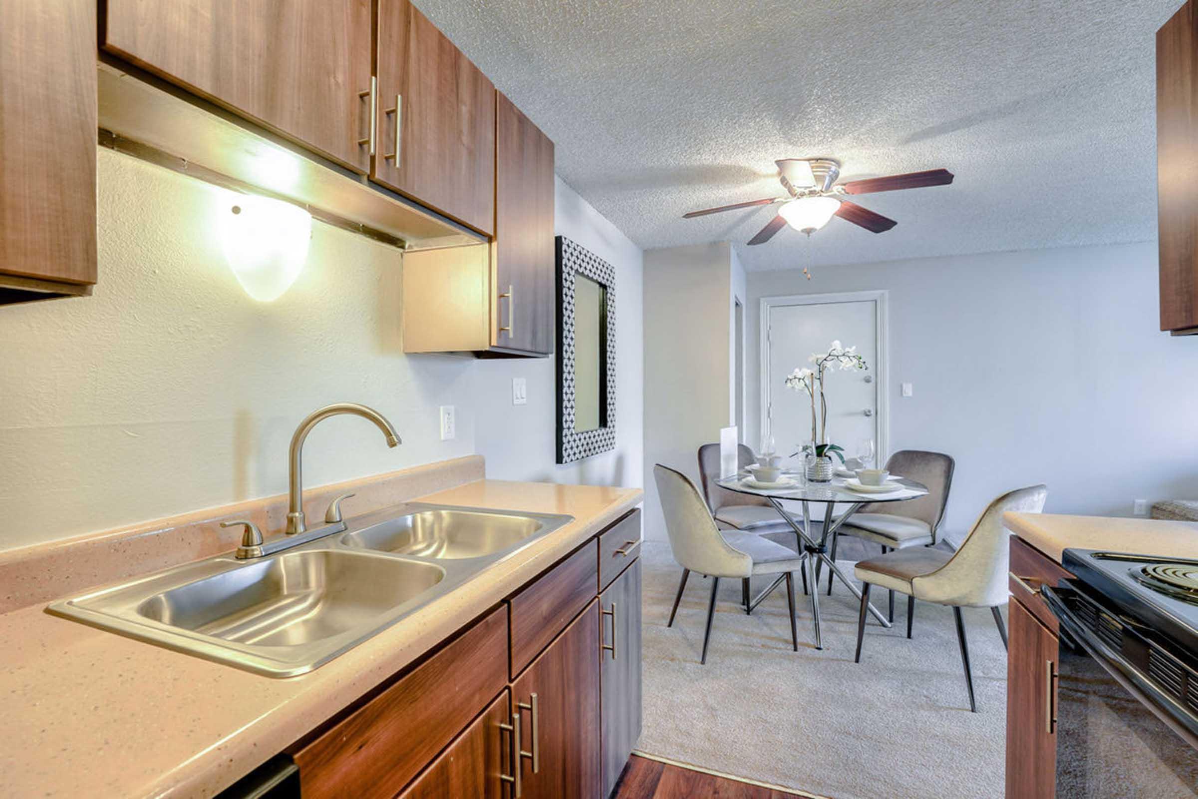 A modern kitchen featuring wooden cabinets, a stainless steel double sink, and a countertop with appliances. The adjacent dining area includes a round glass table with chairs, elegantly decorated with a vase of flowers. A ceiling fan and soft lighting enhance the inviting ambiance.