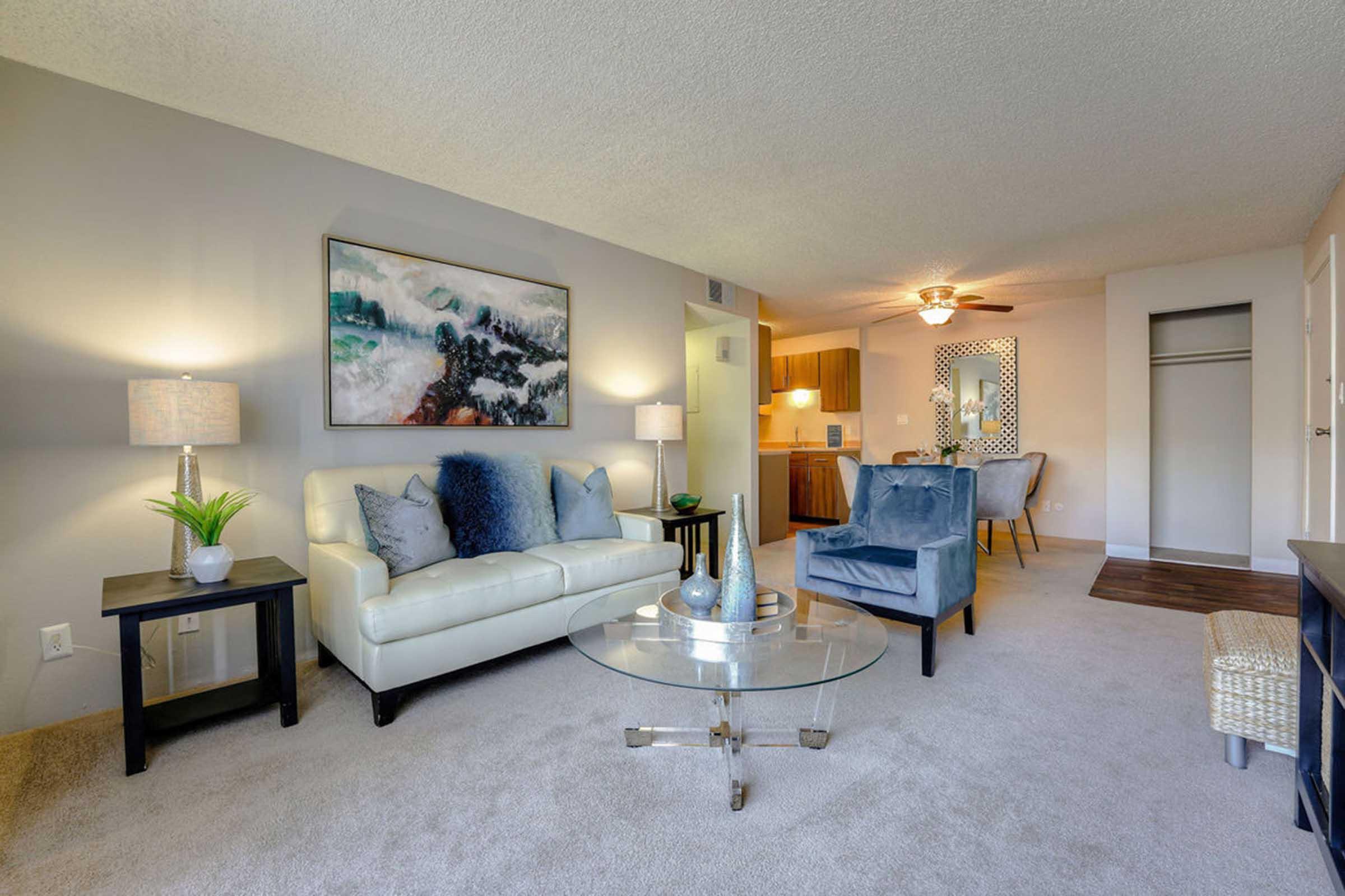 A cozy living room featuring a white sofa with blue and gray decorative pillows, a glass coffee table, and two side tables with lamps. A piece of abstract artwork hangs on the wall, and there's a blue armchair nearby. Light-colored walls and carpet create a warm atmosphere, with a glimpse of a kitchen area in the background.