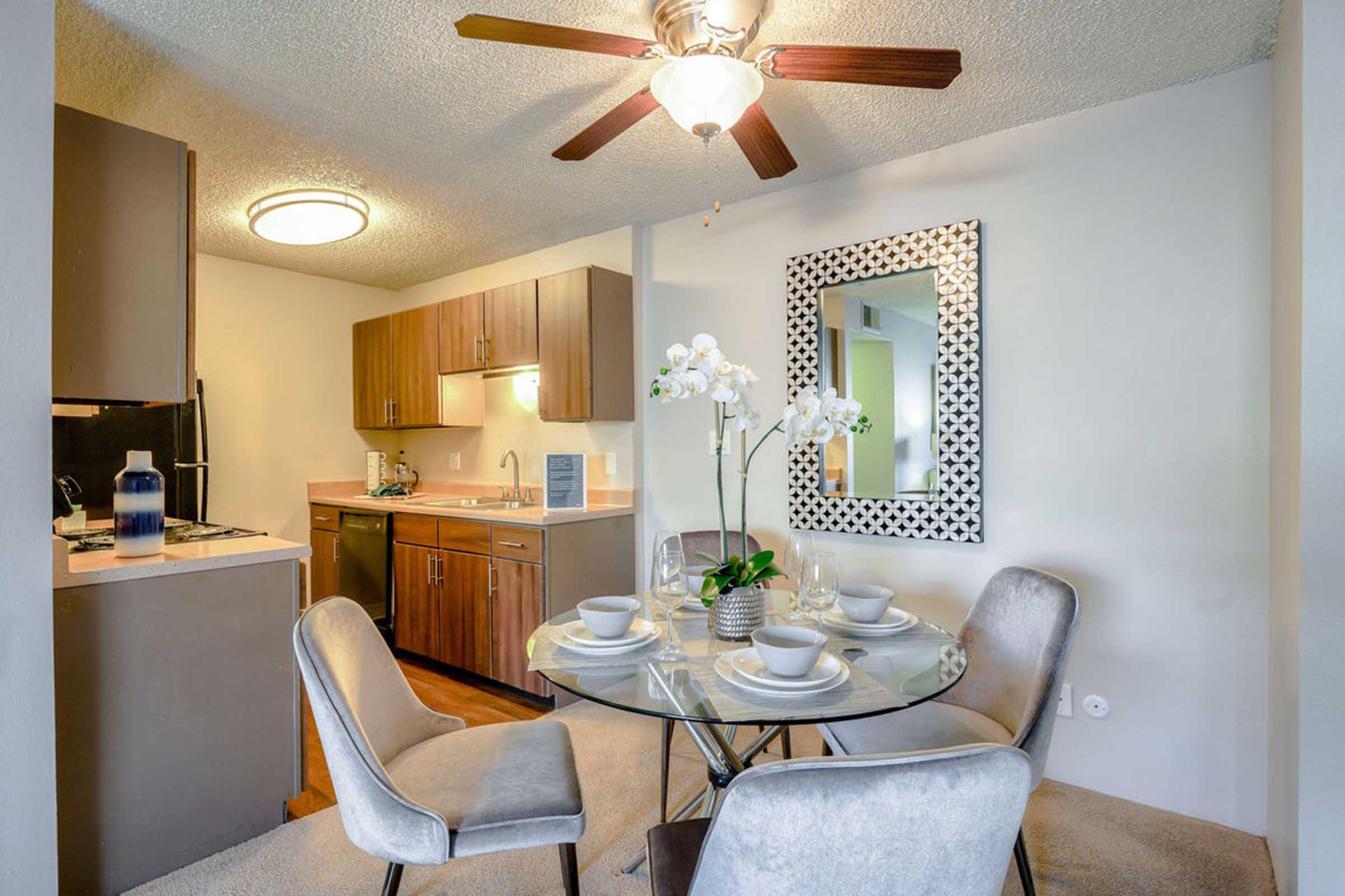 A bright dining area featuring a round glass dining table set for four, with gray upholstered chairs. Behind it, a modern kitchen with wooden cabinets and a small countertop. A ceiling fan is above, and a decorative mirror with a patterned frame hangs on the wall. A potted orchid adds a touch of elegance to the setting.