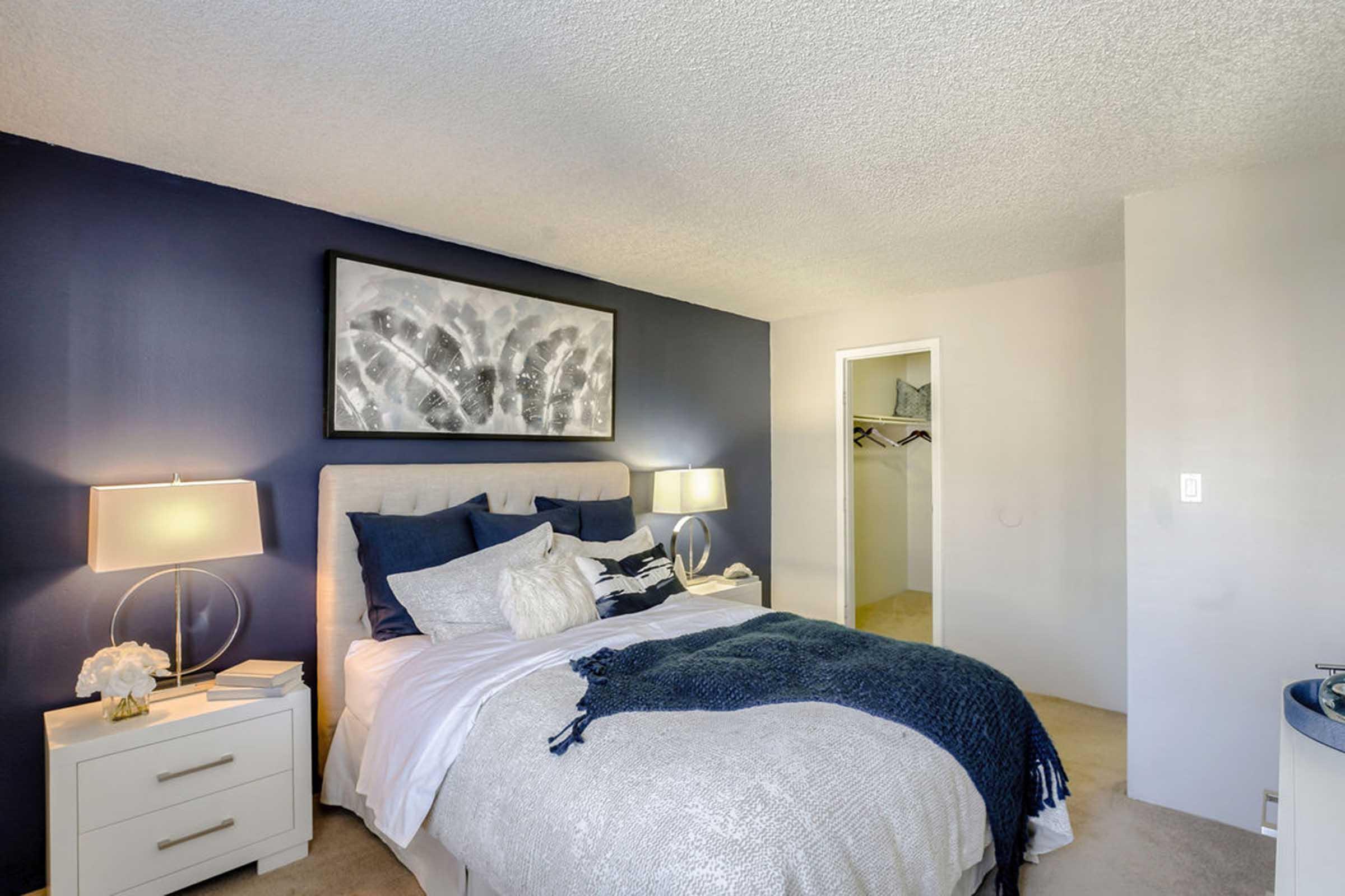 A cozy bedroom featuring a bed with white and blue bedding, two bedside lamps on white nightstands, and a large piece of abstract wall art. The walls are painted in a deep blue, and there is a closet visible in the background. The room has soft lighting, creating a warm and inviting atmosphere.