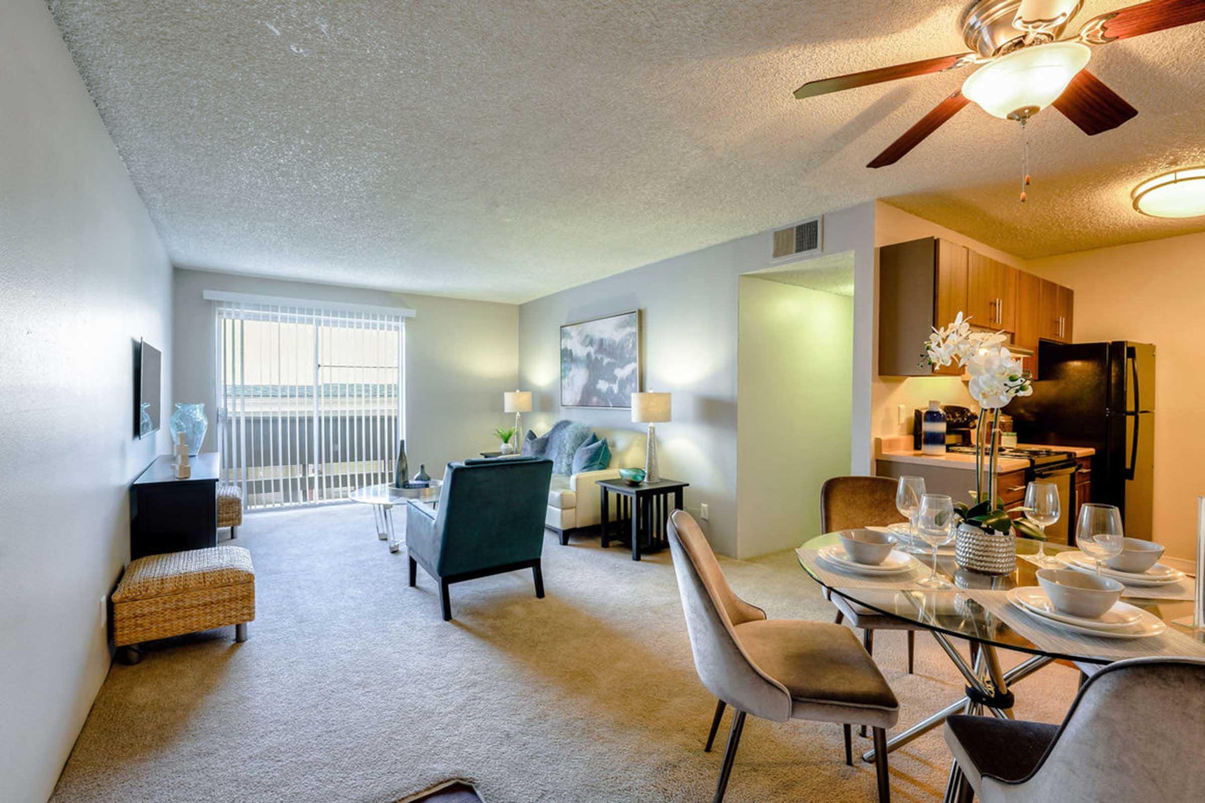 A modern living room and kitchen area featuring a cozy couch, two chairs, a dining table set for four, and a stylish décor. Large windows allow natural light in, with a view of a balcony. The kitchen includes dark cabinetry and appliances, and the space has neutral-colored walls and carpet.
