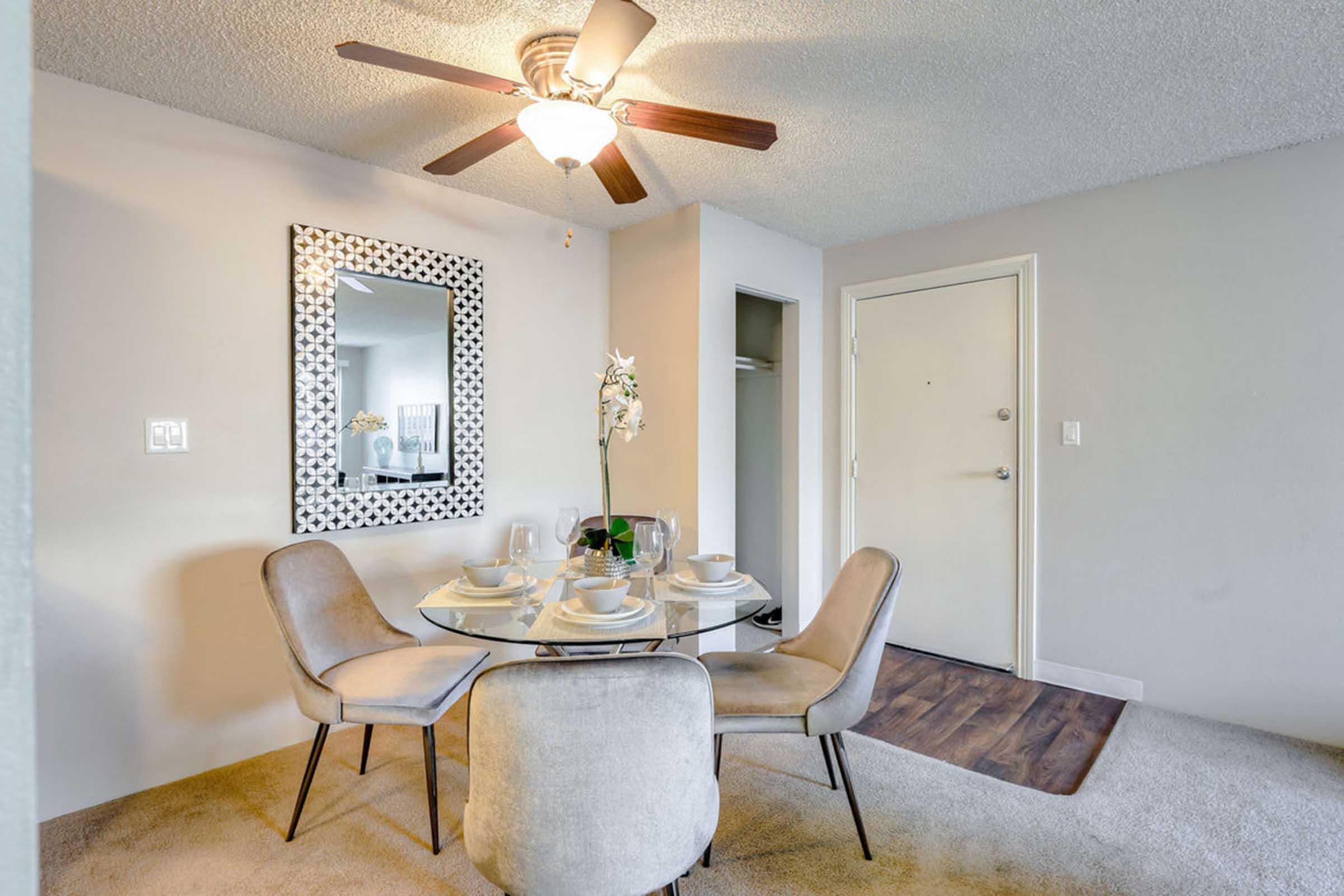A cozy dining area featuring a round glass table set for four with dishes, a decorative mirror on the wall, and a ceiling fan. The walls are light-colored, and there's an entrance door visible on the right. The floor has a warm wooden finish, complementing the neutral-toned furniture.