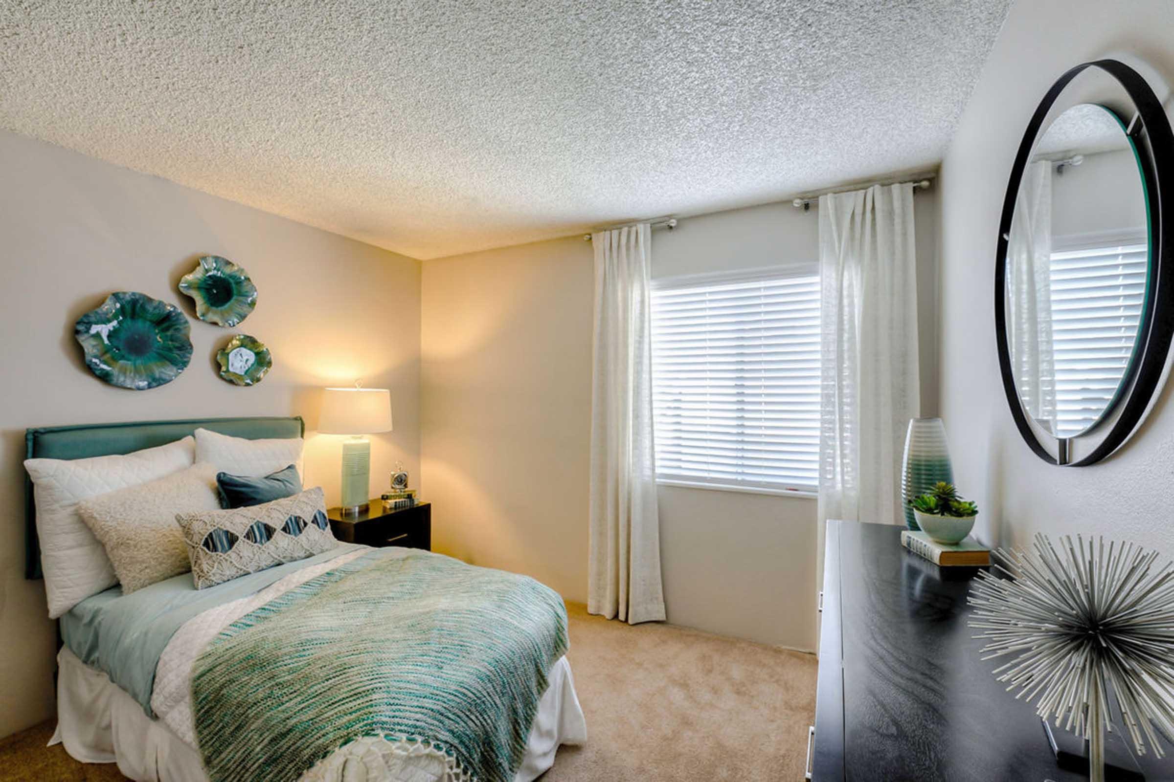 A beautifully decorated bedroom featuring a bed with teal and white bedding, accent pillows, and a nightstand with a lamp. Decorative wall art in teal hues is displayed above the bed. Natural light filters through sheer curtains covering a window, and a round mirror reflects the room's cozy ambiance.