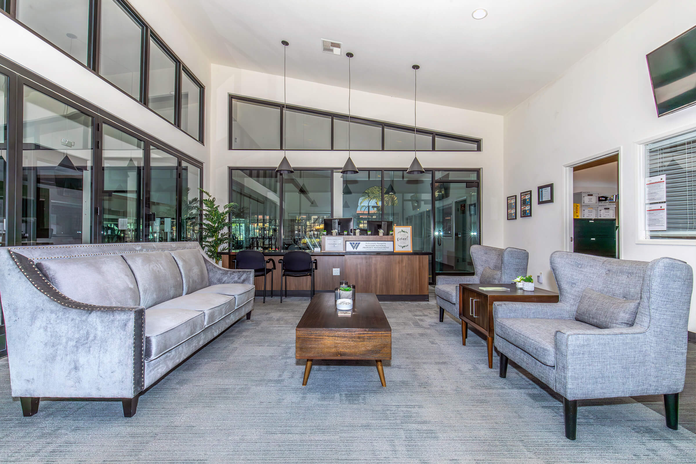 A modern lobby area featuring a grey upholstered sofa, two light grey chairs, a wooden coffee table, and a reception desk in the background. Large windows allow natural light to fill the space, complemented by potted plants and stylish pendant lighting, creating a welcoming atmosphere.
