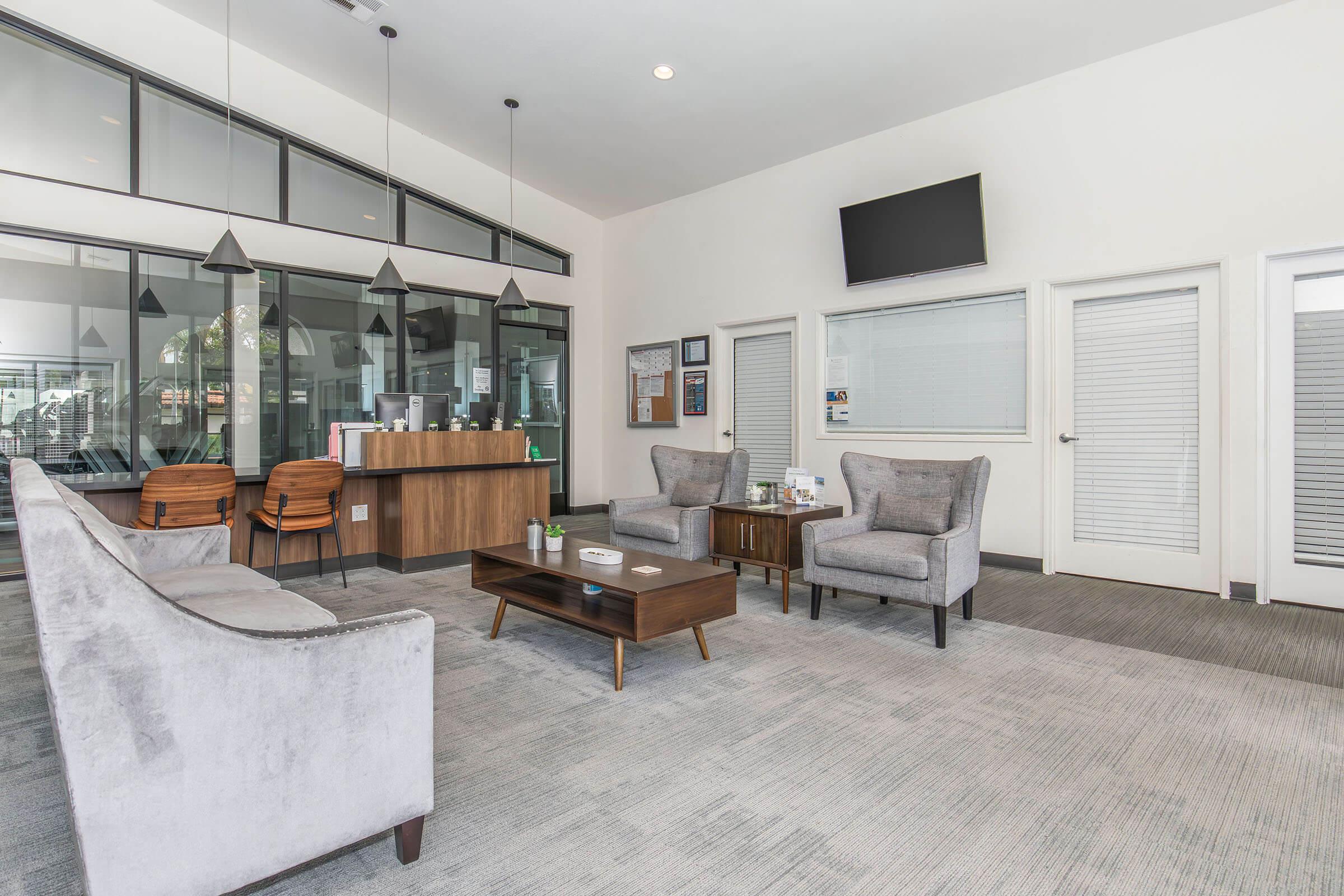 A modern waiting area featuring gray upholstered chairs, a wooden coffee table, and a reception desk in the background. The space has large windows allowing natural light, pendant lights hanging from the ceiling, and a flat-screen TV mounted on the wall. The decor is clean and inviting.