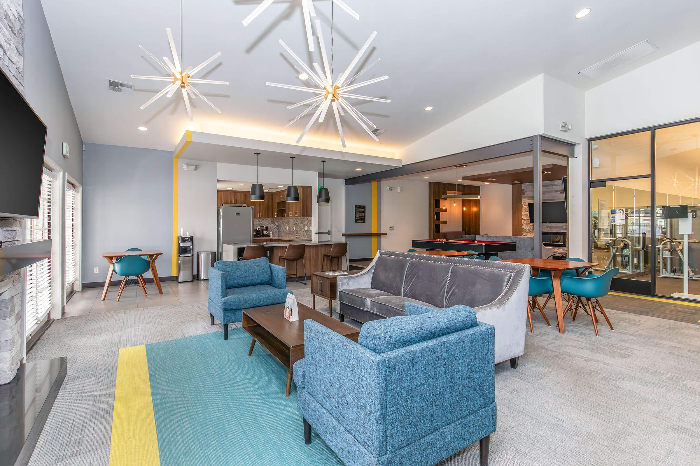 A modern lounge area featuring stylish furniture, including two blue chairs and a gray couch, arranged around a coffee table. Decorative light fixtures hang from the ceiling, and there's a vibrant area rug on the floor. In the background, a kitchen and fitness area are visible through glass partitions.