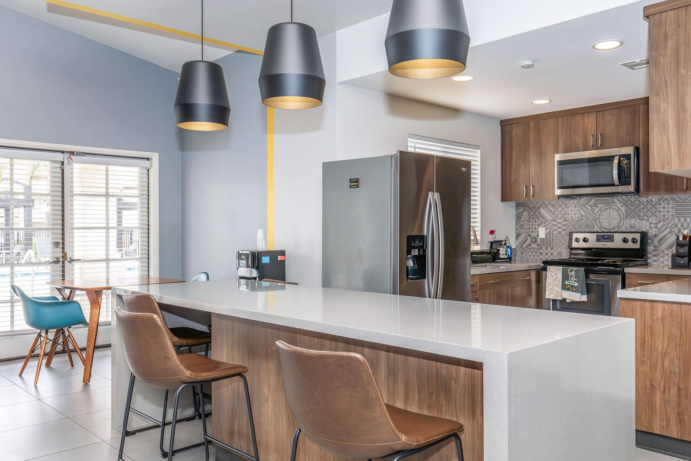 Modern kitchen featuring a large island with gray countertops and wooden cabinetry. Three pendant lights hang above the island, which has two brown stools. The kitchen includes stainless steel appliances and a patterned backsplash, with large windows providing natural light.
