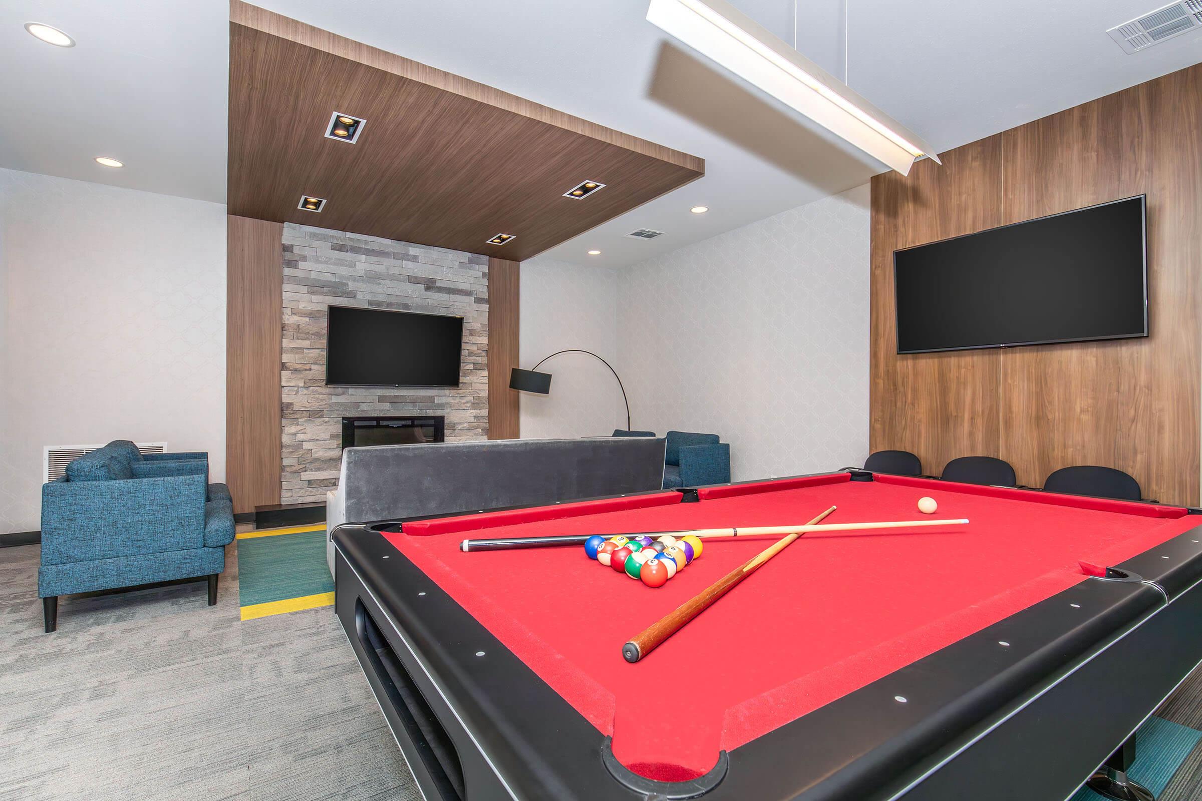 A well-lit game room featuring a red pool table with balls and cues ready for play. In the background, there are cozy seating areas with blue chairs, a modern fireplace, and two wall-mounted televisions. The room has a contemporary design with wooden and textured walls, creating a relaxed atmosphere for leisure activities.