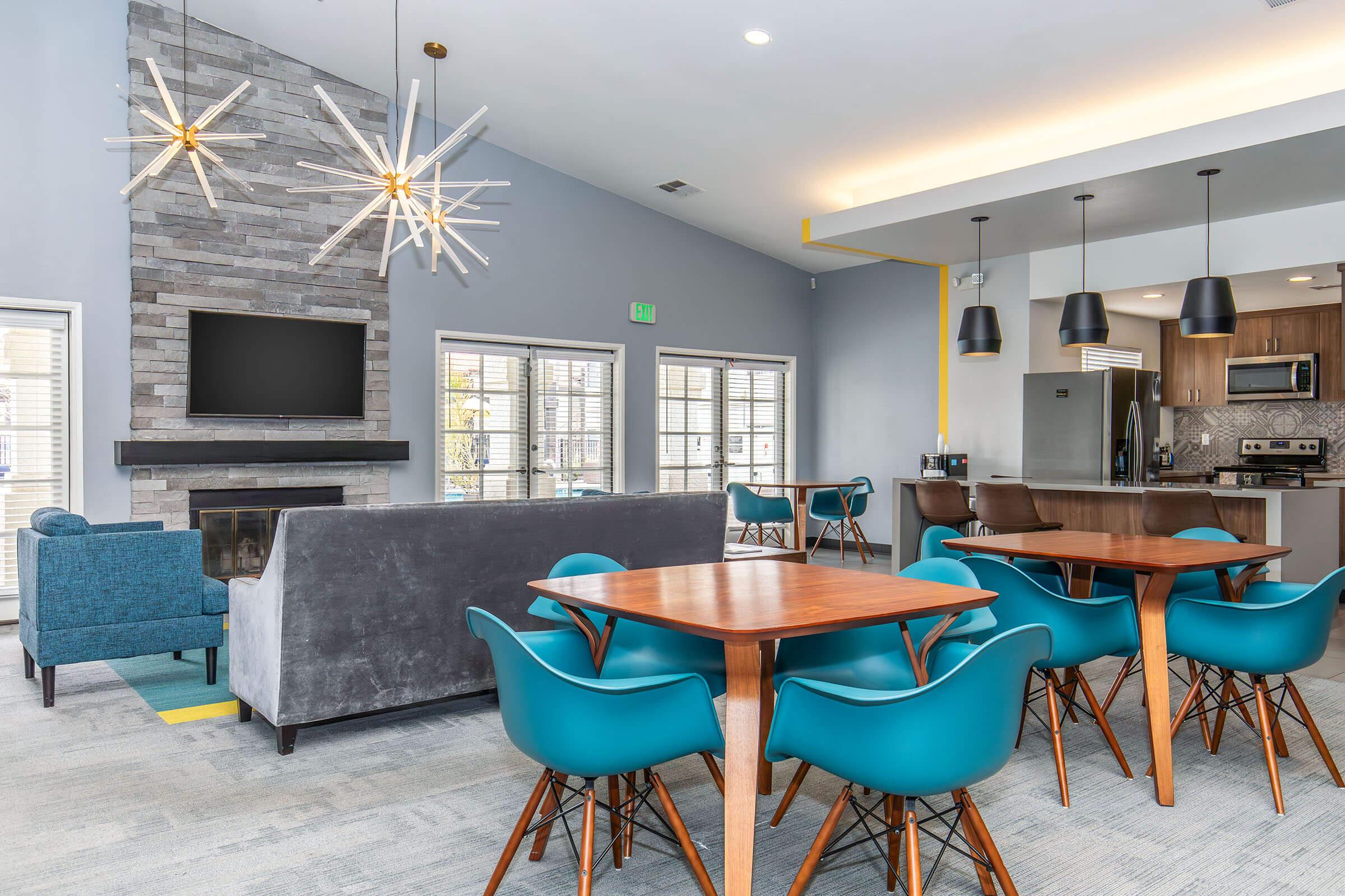 A modern communal living space featuring a cozy seating area with a gray couch and blue chairs, a stone fireplace with a mounted TV, and several wooden tables with blue chairs. Large windows allow natural light to fill the room, creating a welcoming atmosphere.
