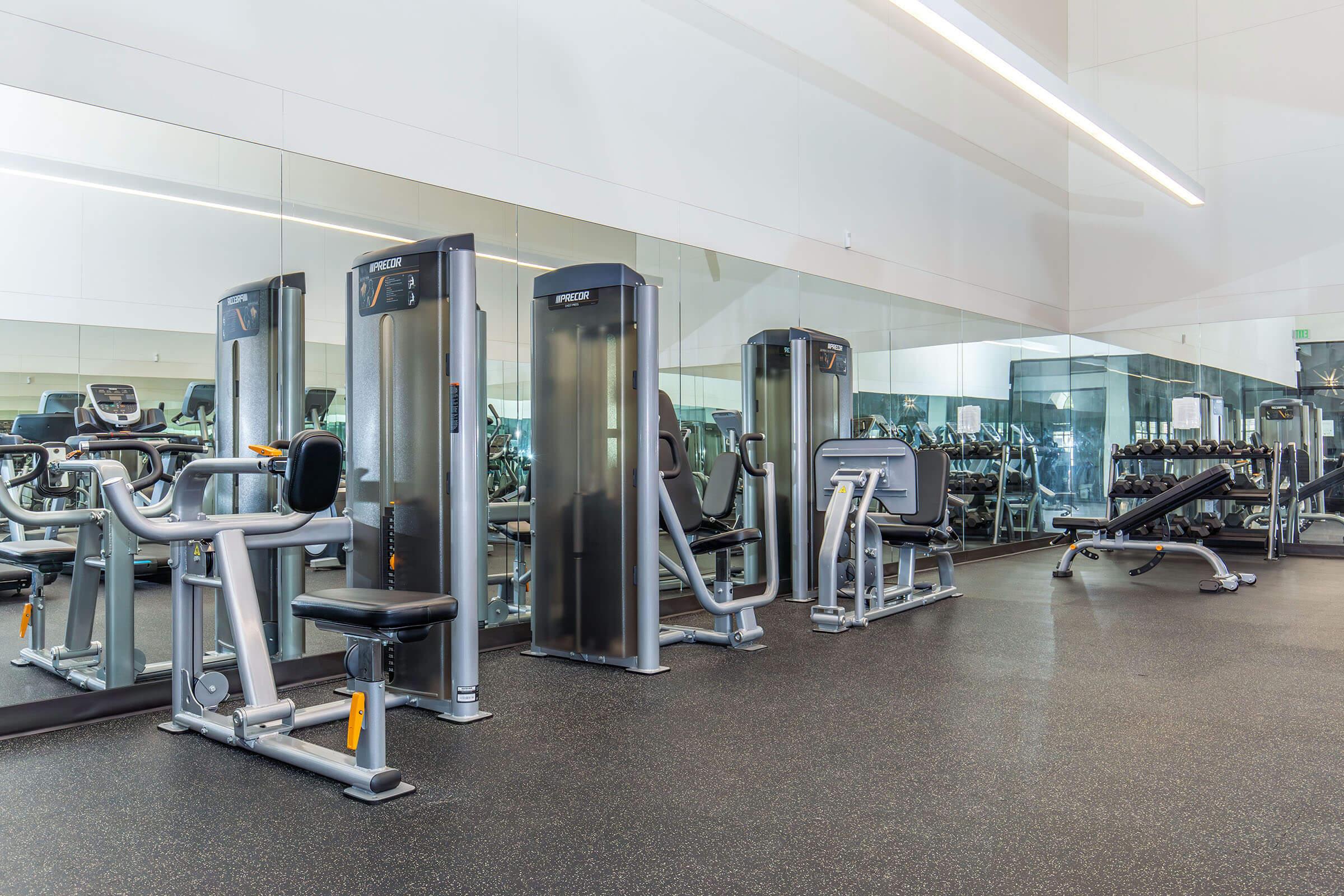 A modern gym interior featuring various exercise equipment, including strength training machines and a bench, with a large mirror reflecting the space. The flooring is dark and rubberized, providing a workout-friendly surface, and there are additional gym machines visible in the background.