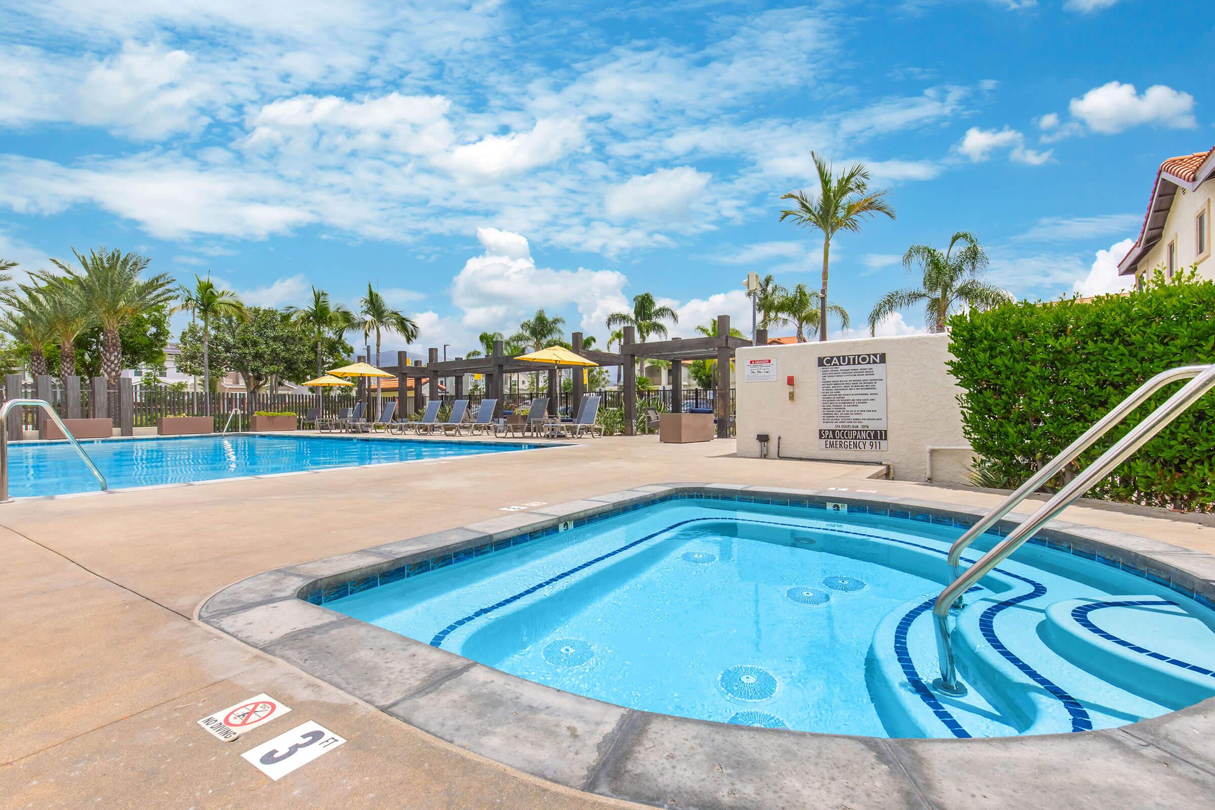 A clear blue swimming pool with a hot tub in the foreground. Sun loungers are arranged in the background under yellow umbrellas. Lush palm trees and a bright blue sky with scattered clouds create a vibrant atmosphere, indicating a warm and inviting outdoor space. Safety signs are visible nearby.