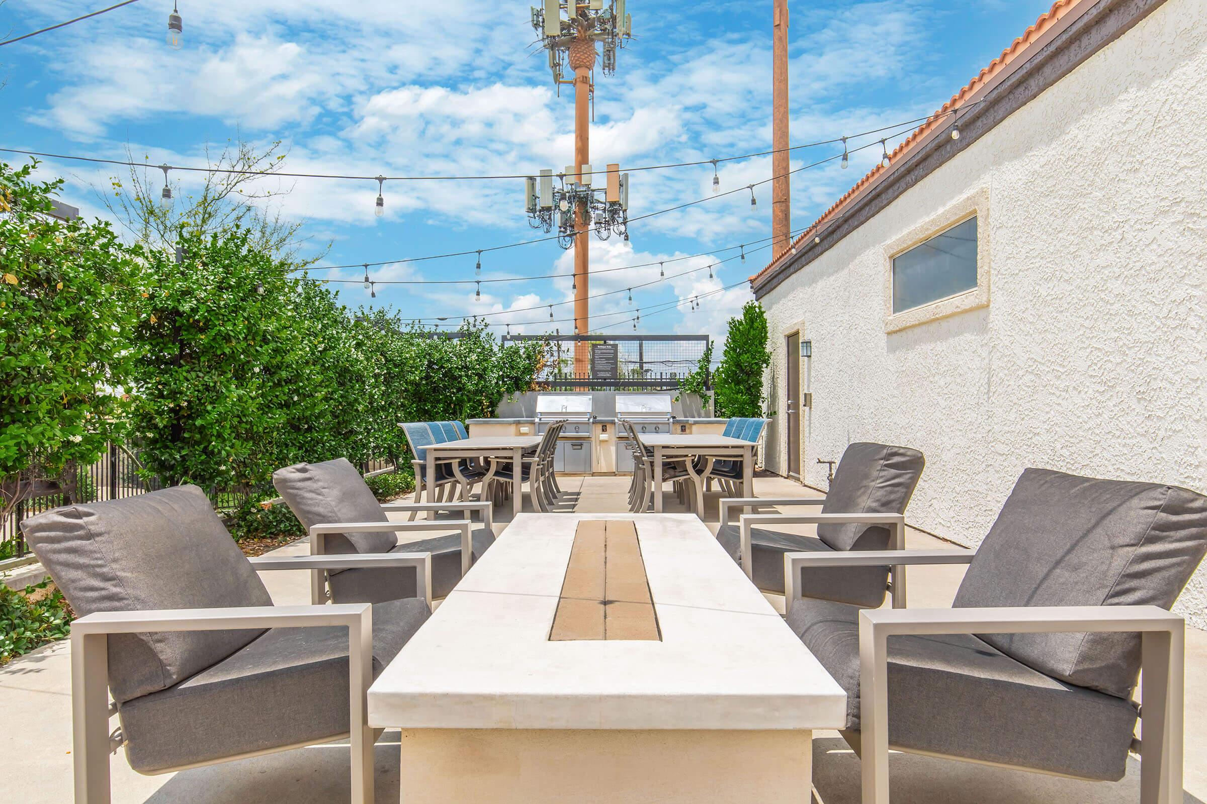Outdoor seating area featuring a long table surrounded by several gray chairs. The background includes a white wall, green foliage, and cell towers. Bright blue sky with scattered clouds overhead, and string lights hanging above the seating area.