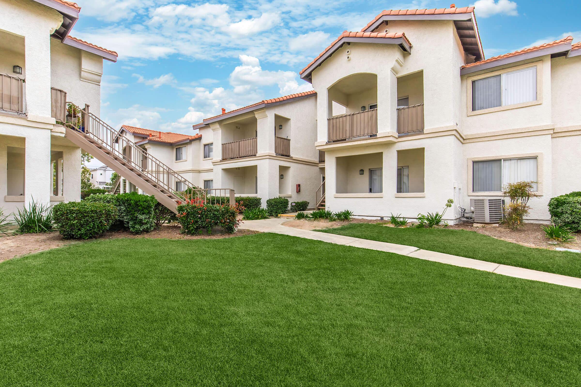 A well-maintained apartment complex featuring two-story buildings with white exteriors and terracotta roofs. The foreground includes a grassy lawn and a walkway leading to a staircase. The scene is set under a partly cloudy blue sky, creating a welcoming atmosphere.