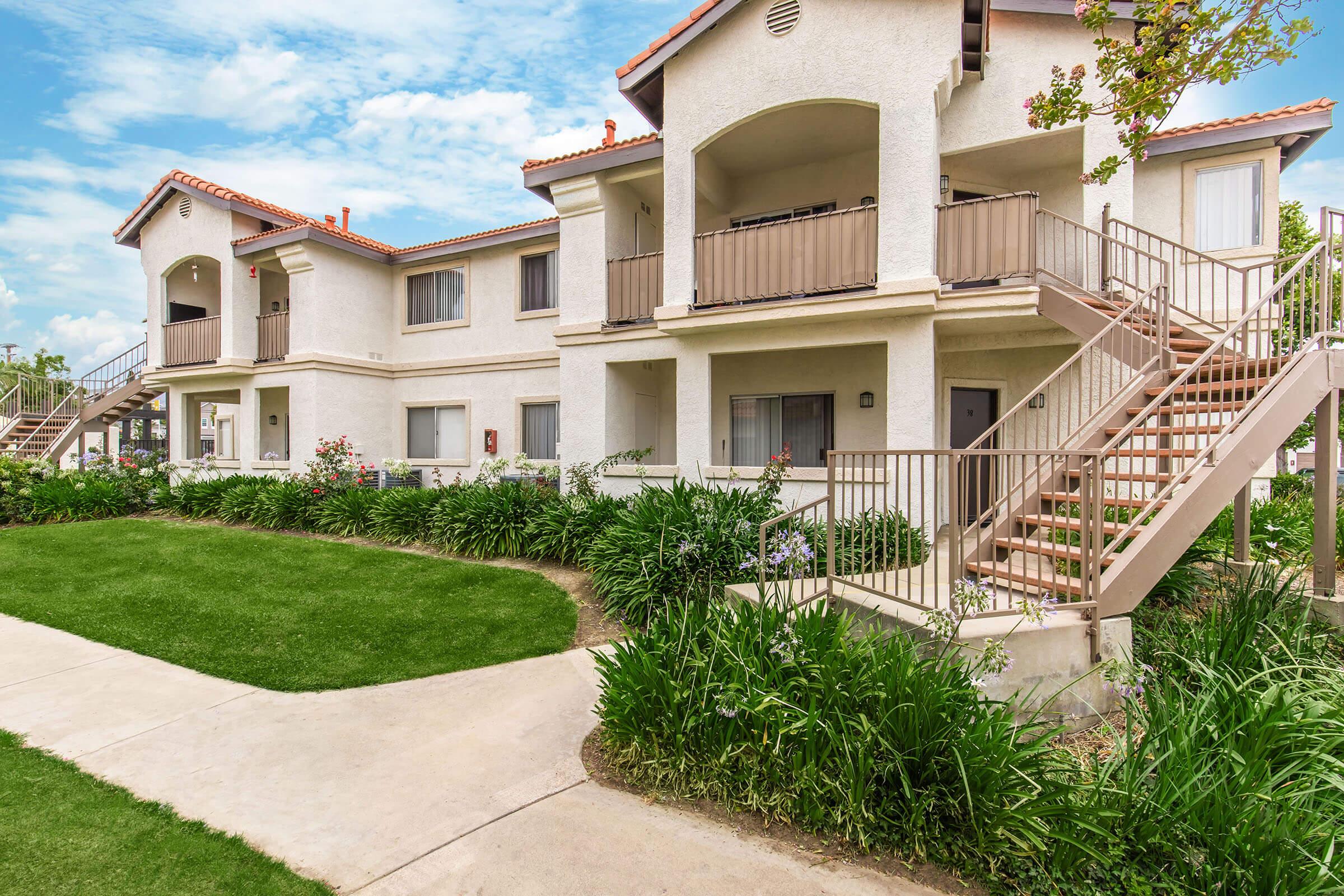 Two-story apartment building with a light-colored exterior and a red-tiled roof, surrounded by well-maintained green lawns and shrubbery. A staircase leads to the second floor, where several units feature balconies. The scene is bright and inviting, under a partly cloudy blue sky.