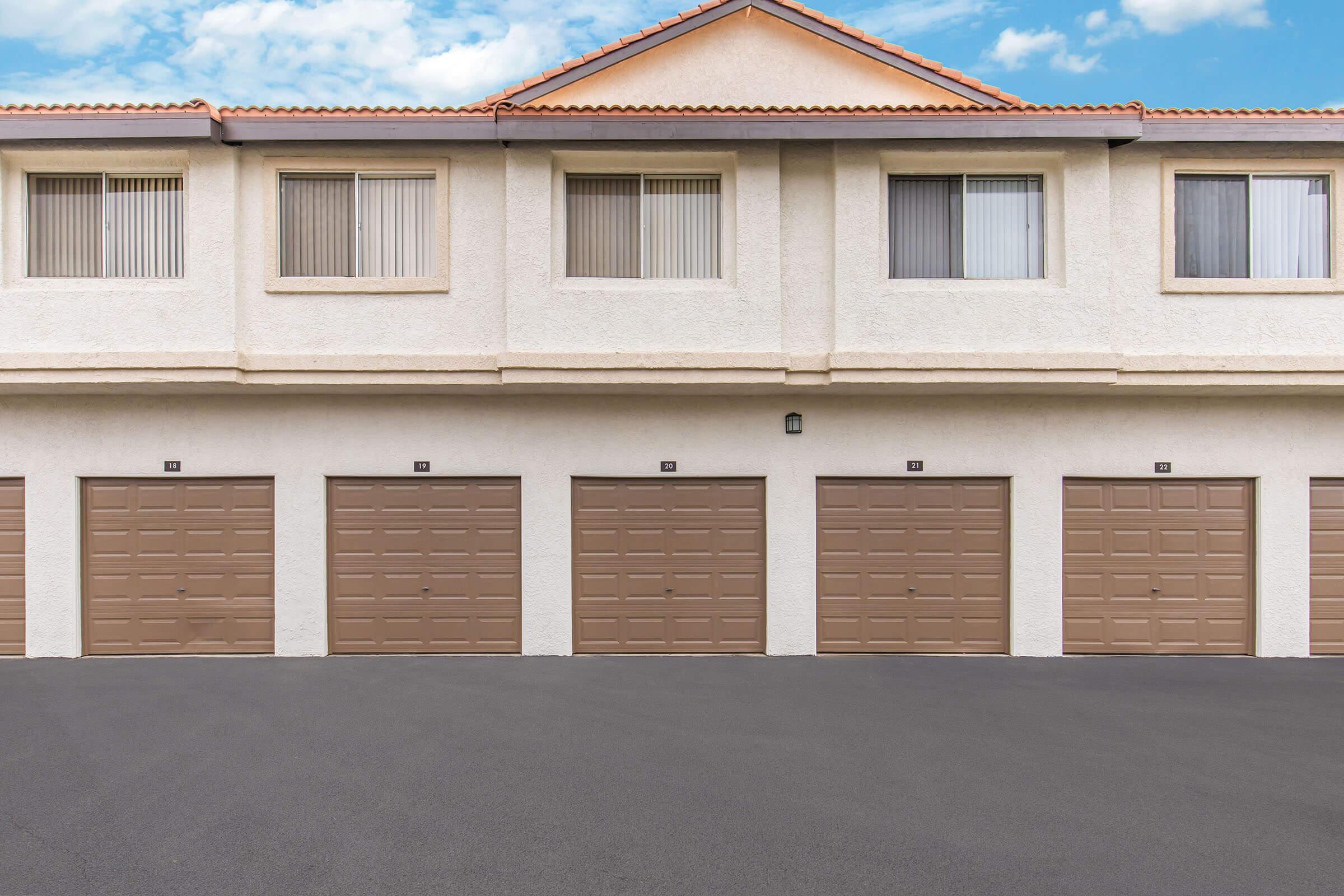 A row of four tan garage doors beneath a light-colored apartment building. The facade features a tile roof and several windows with vertical blinds. The sky above is mostly clear with a few clouds, and the ground is paved with dark asphalt.