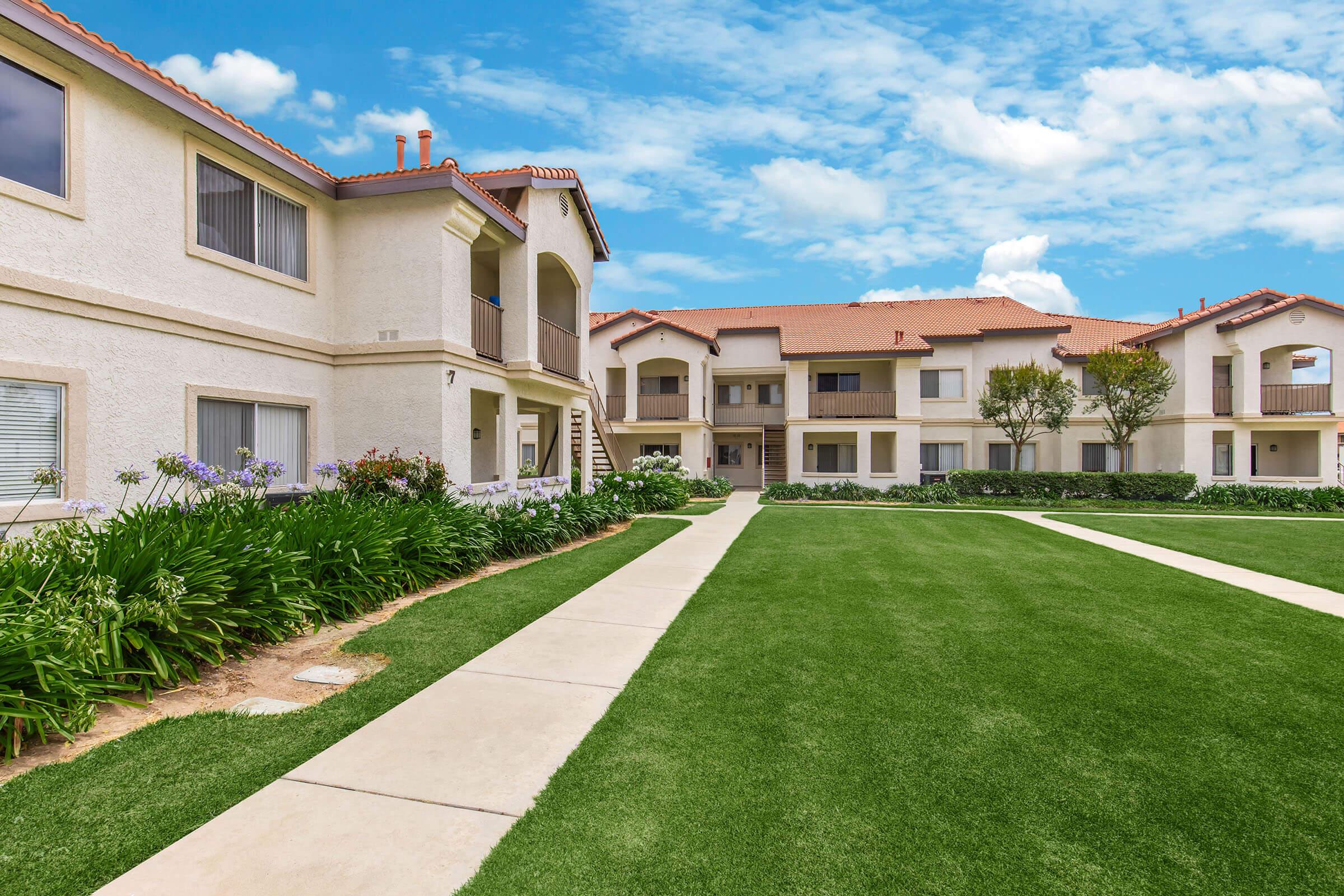 A well-maintained residential complex featuring two-story buildings with balconies, surrounded by green lawns and pathways. The sky is bright with scattered clouds, creating a pleasant ambiance. Flower beds line the walkways, enhancing the appealing outdoor space.