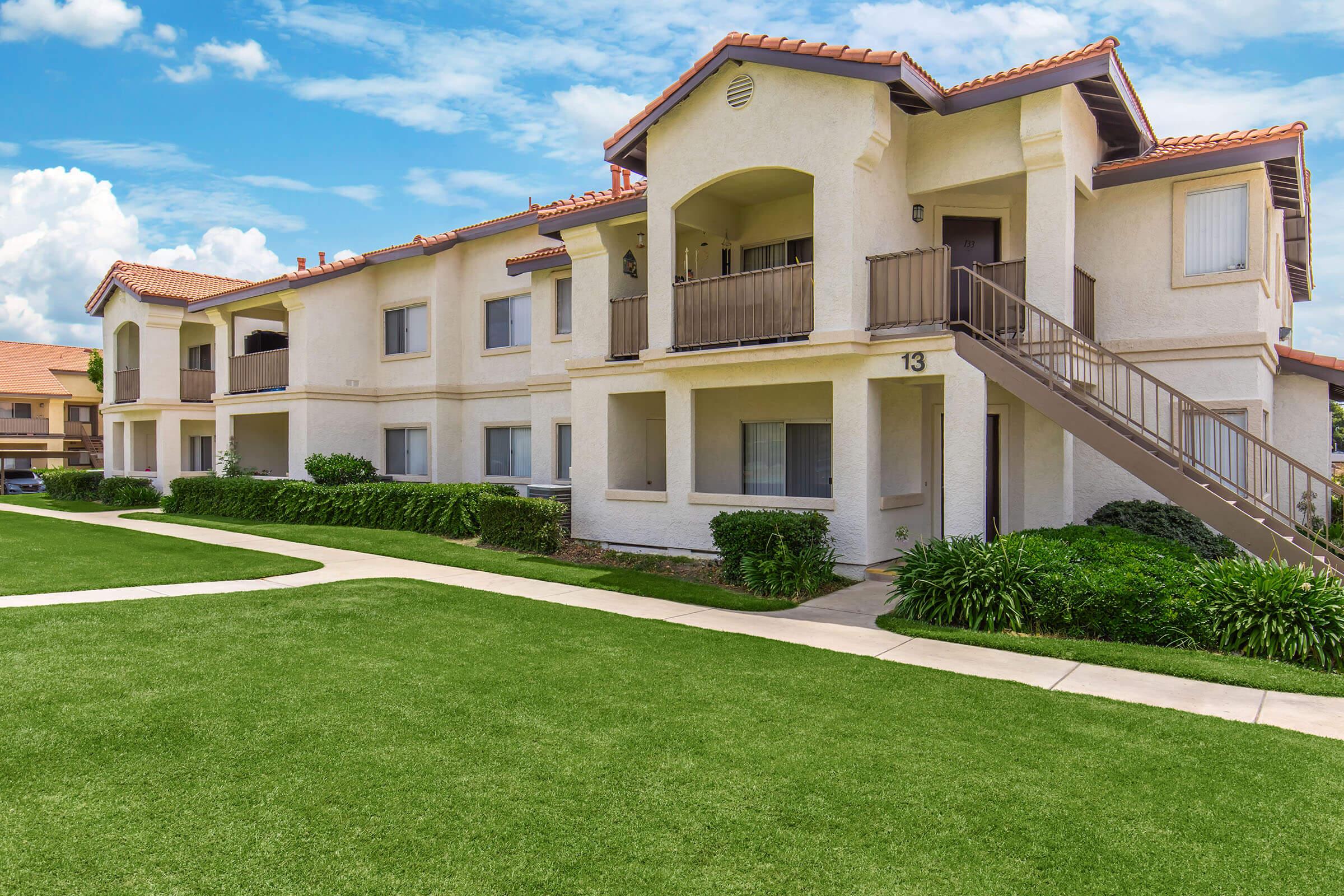 A modern two-story apartment building with a light-colored exterior and tiled roofs. It features multiple balconies and large windows, surrounded by neatly trimmed grass and landscaping. The sky is blue with fluffy white clouds, creating a bright and inviting atmosphere.