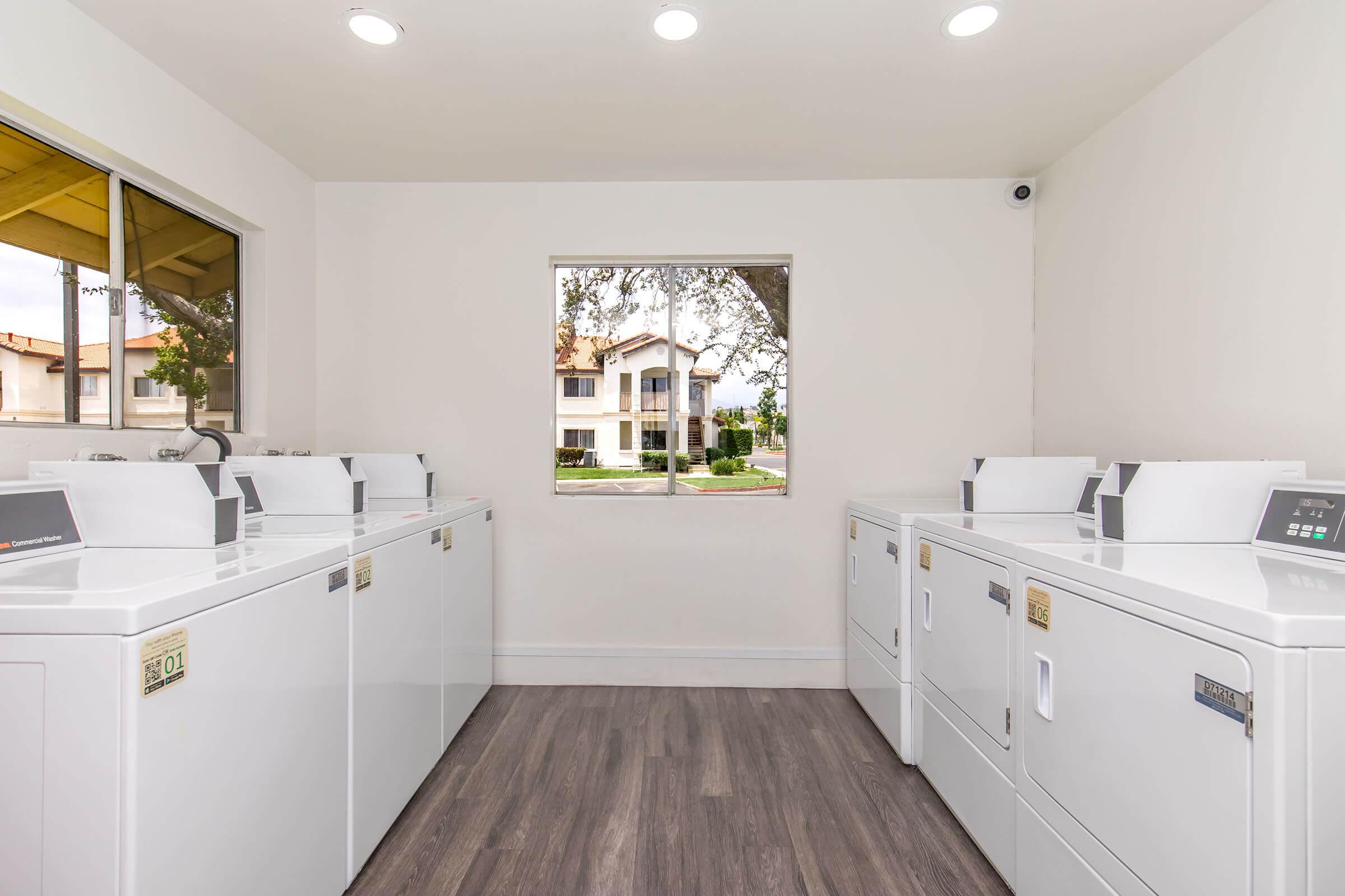 A bright and clean laundry room featuring several white washing machines and dryers arranged against the wall. A window offers a view of a residential area outside, with a well-maintained lawn and buildings visible. The ceiling has recessed lighting, enhancing the room's light and airy atmosphere.